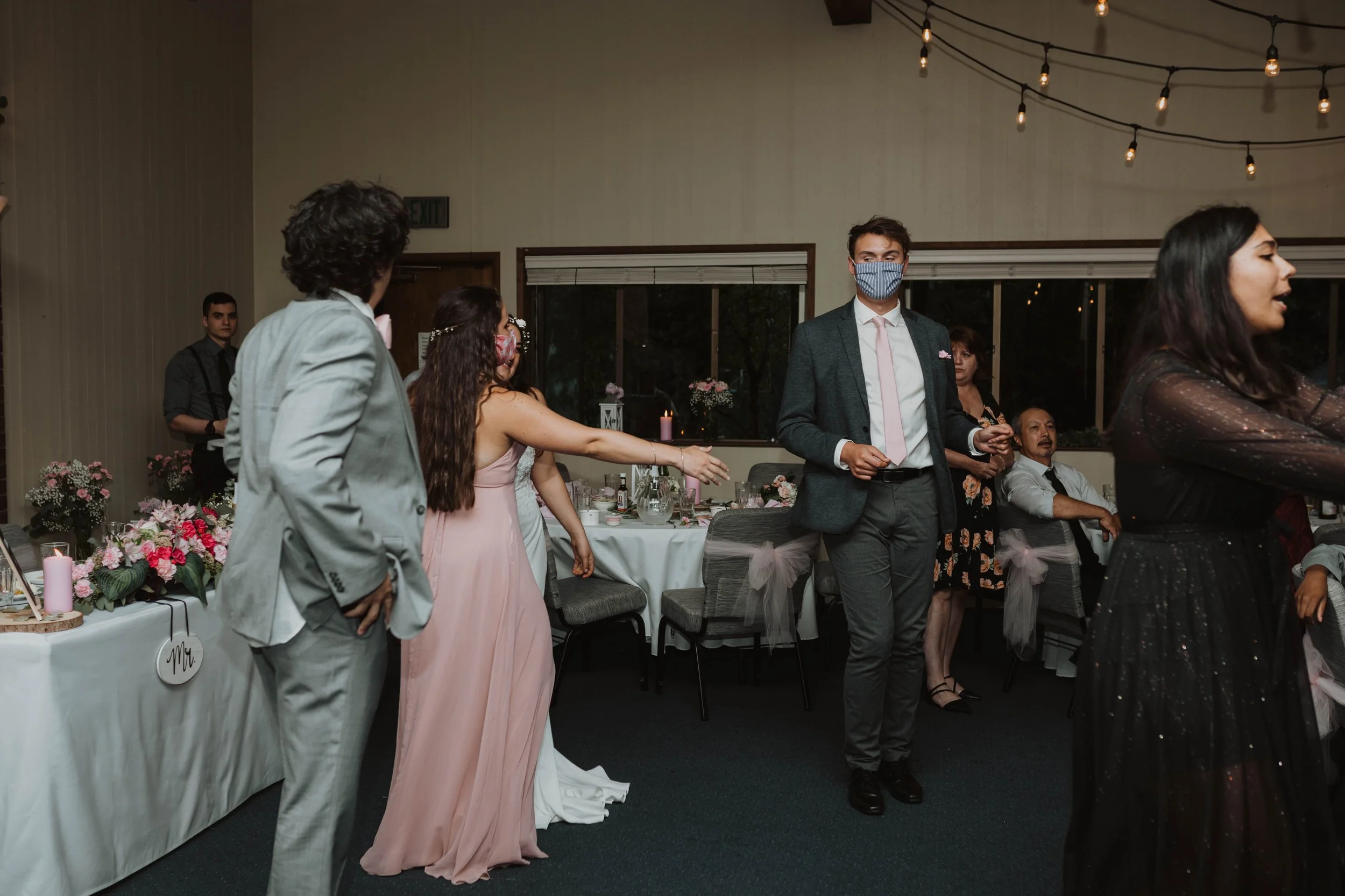 Guests wearing dresses and suits, some with masks, dancing at a wedding reception in a decorated indoor venue with hanging lights and floral arrangements. Seattle, WA wedding photography.