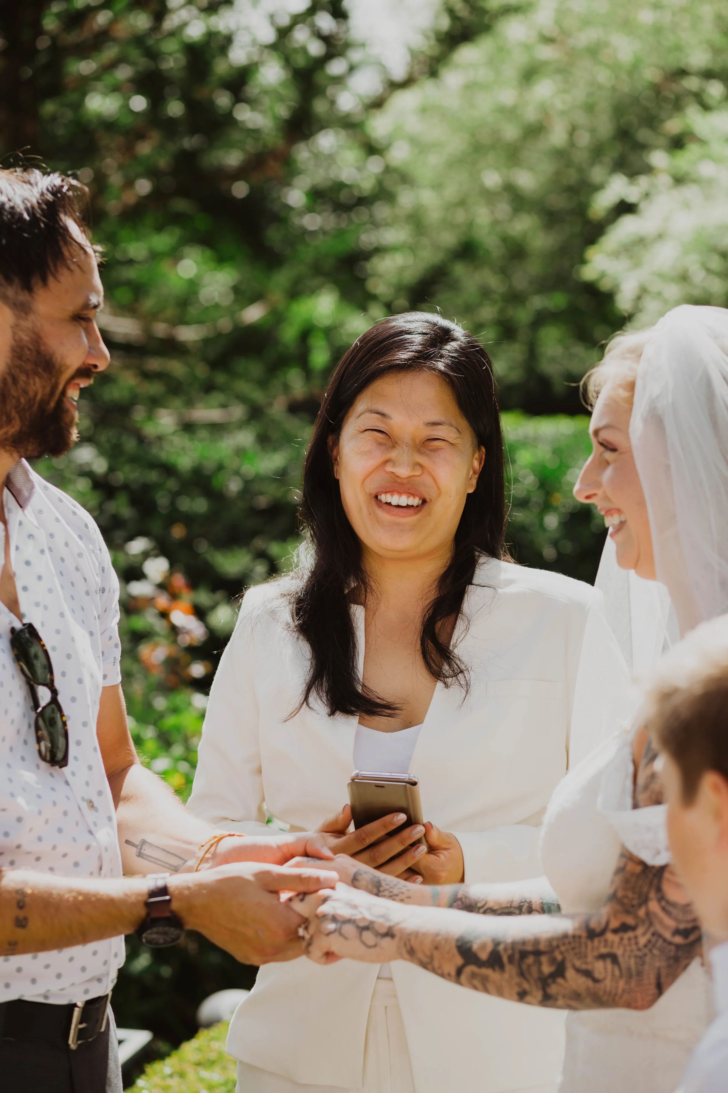 A couple getting married, holding hands, with a woman officiant and another woman in a white dress, outdoors with trees and greenery in the background. Seattle, WA wedding photography.