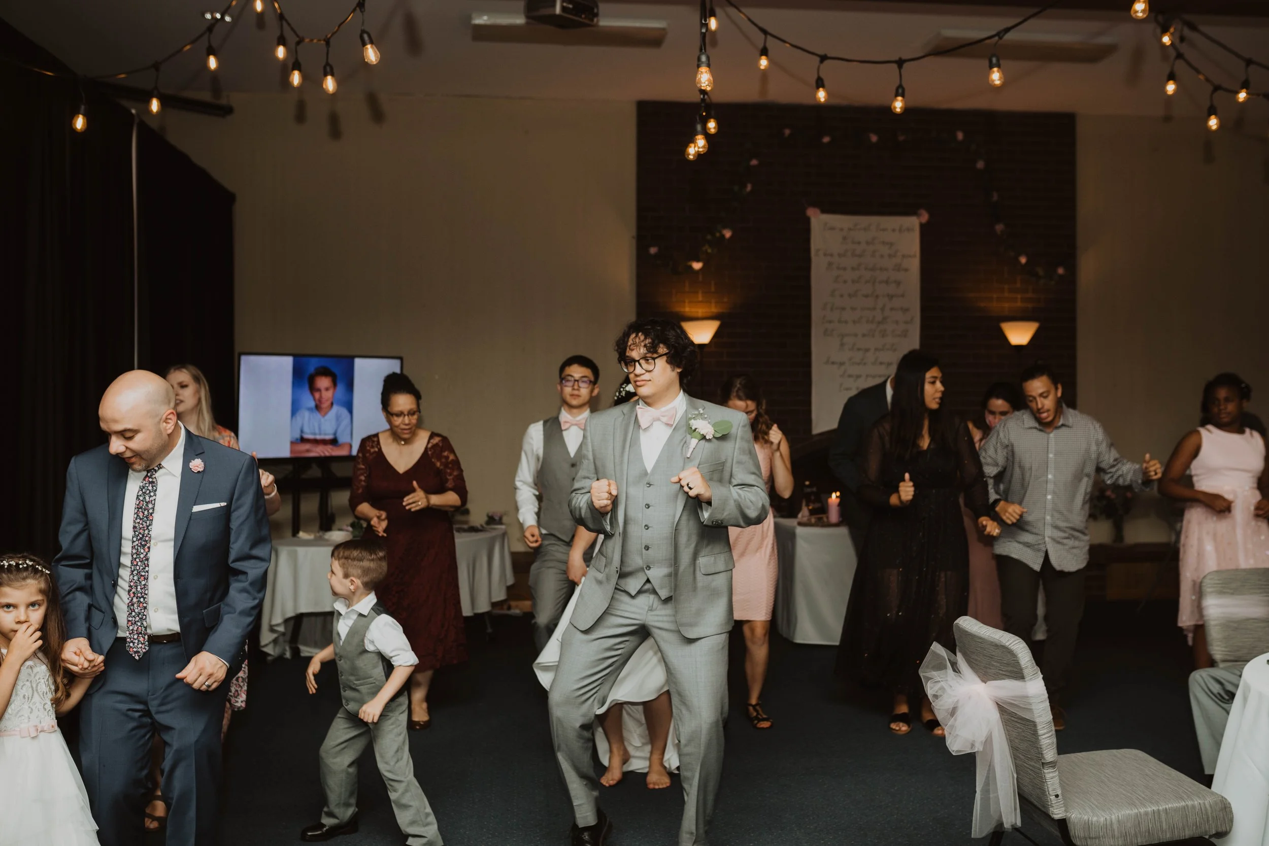 People dancing at a wedding reception in a decorated hall with string lights and a brick wall background. Seattle, WA wedding photography.