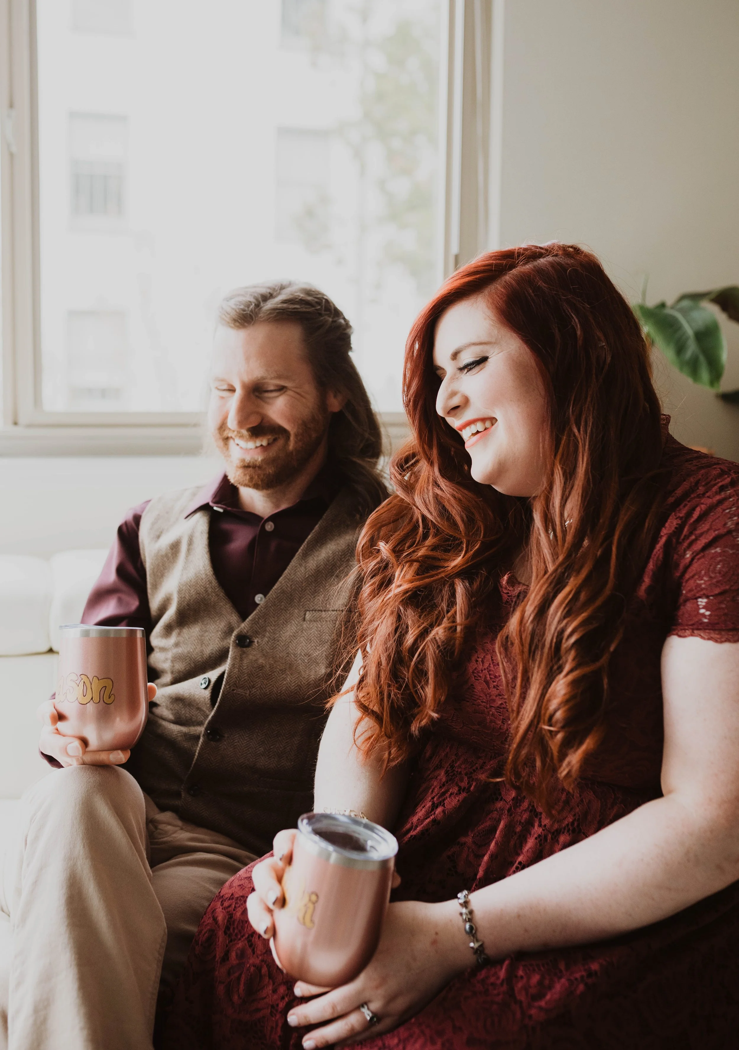 A man and woman sitting on a couch, smiling and holding mugs, with a window behind them. Pioneer Square, Seattle, WA wedding photography.