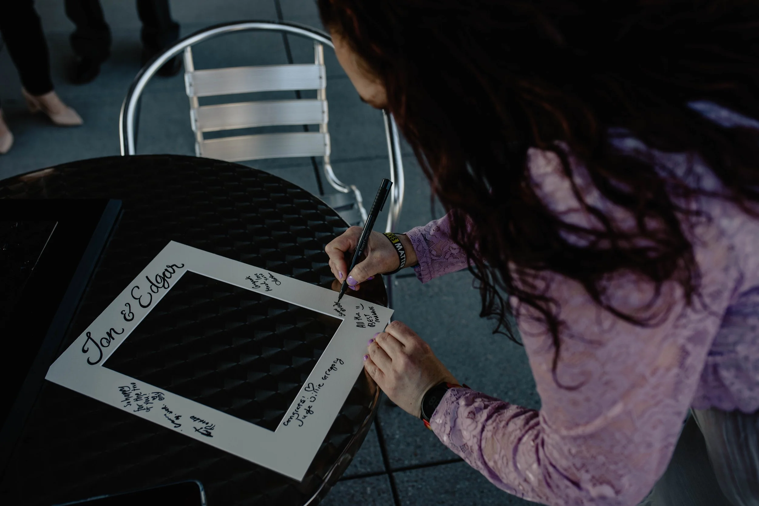 A woman in a pink lace blouse signs a white photo frame with black cursive writing at a table with a black woven surface, with a black folder on the left. Seattle Municipal Courthouse wedding photography.