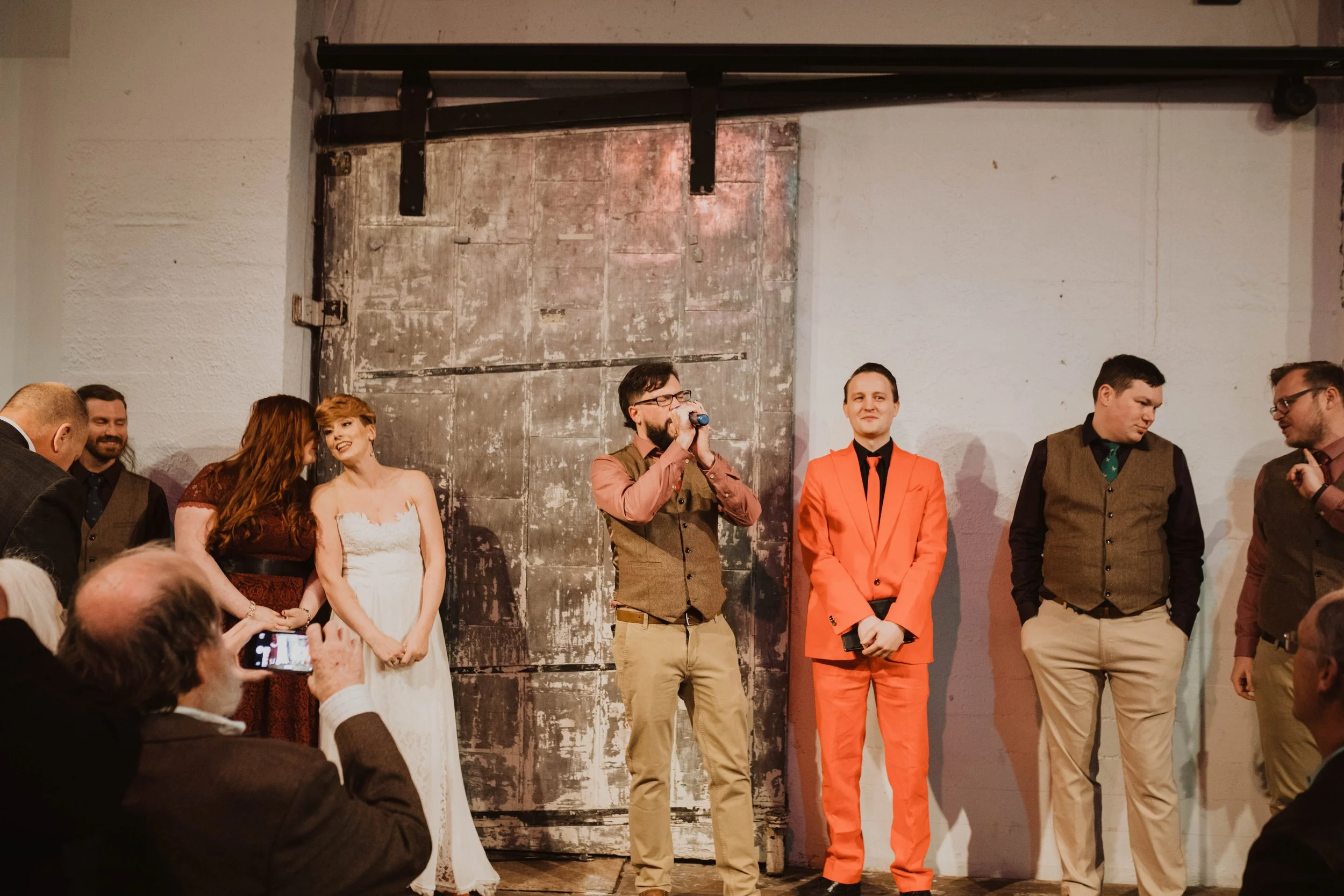 A group of people standing on a stage during a wedding reception. The bride is in a white dress, and the groom is in a bright orange suit. Some individuals are dressed in semi-formal attire. One man is speaking into a microphone, and others are engag