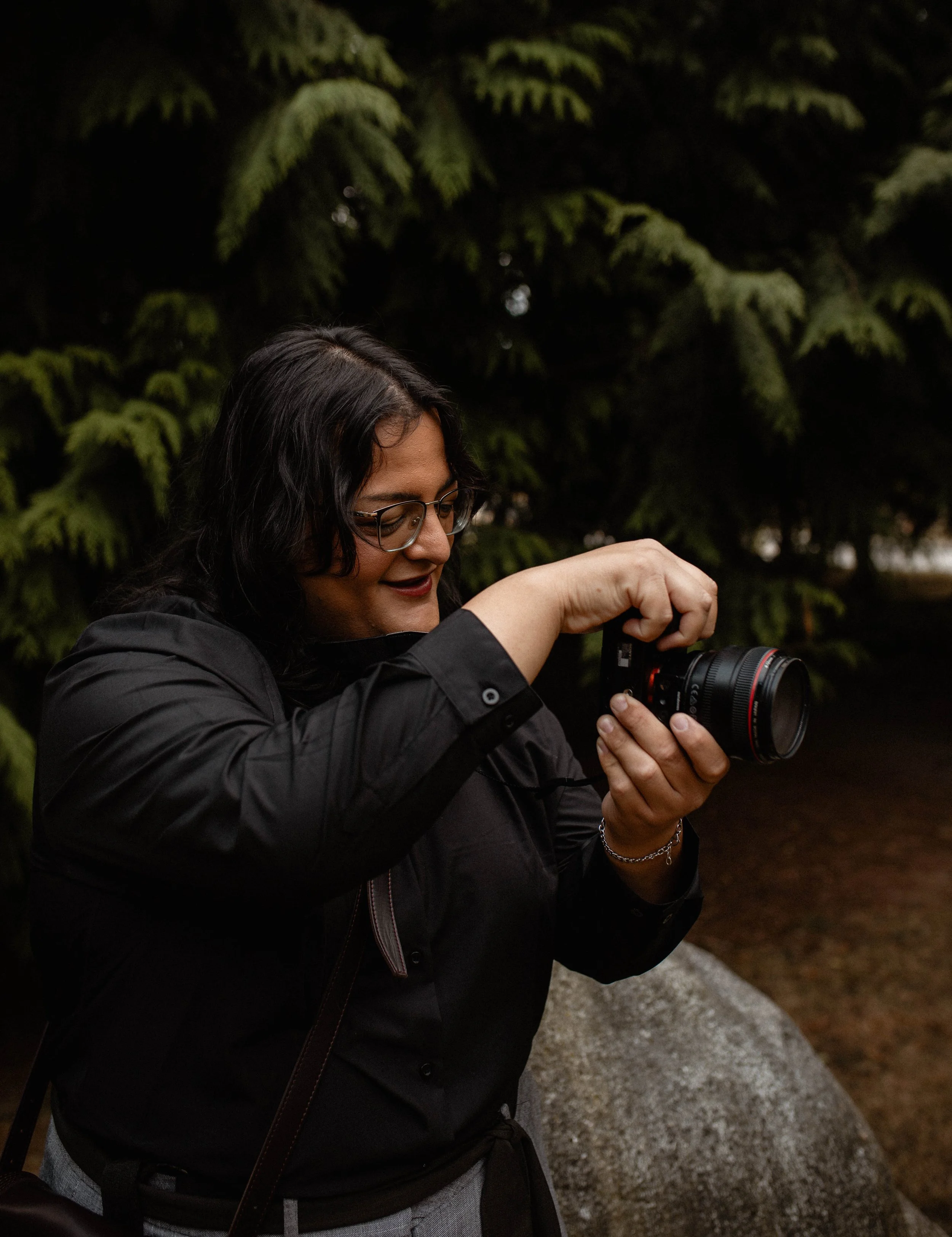 A woman with dark hair and glasses in a black jacket holding a camera outdoors, with a background of green pine trees. Seattle professional head shot photography