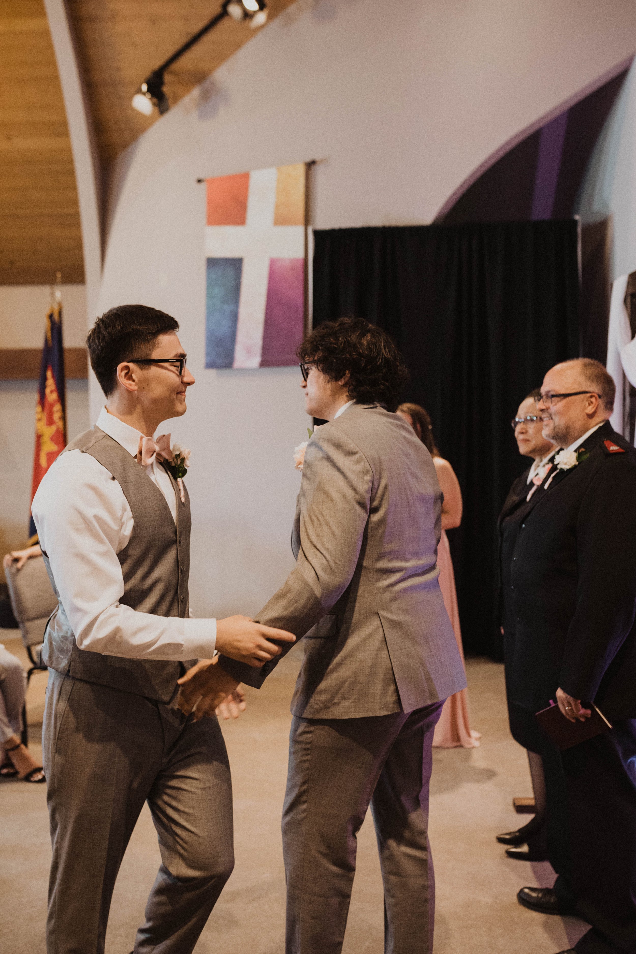 Two men in suits holding hands and looking at each other during a ceremony, with a woman and a man in uniform standing nearby in a decorated indoor venue. Seattle, WA wedding photography.