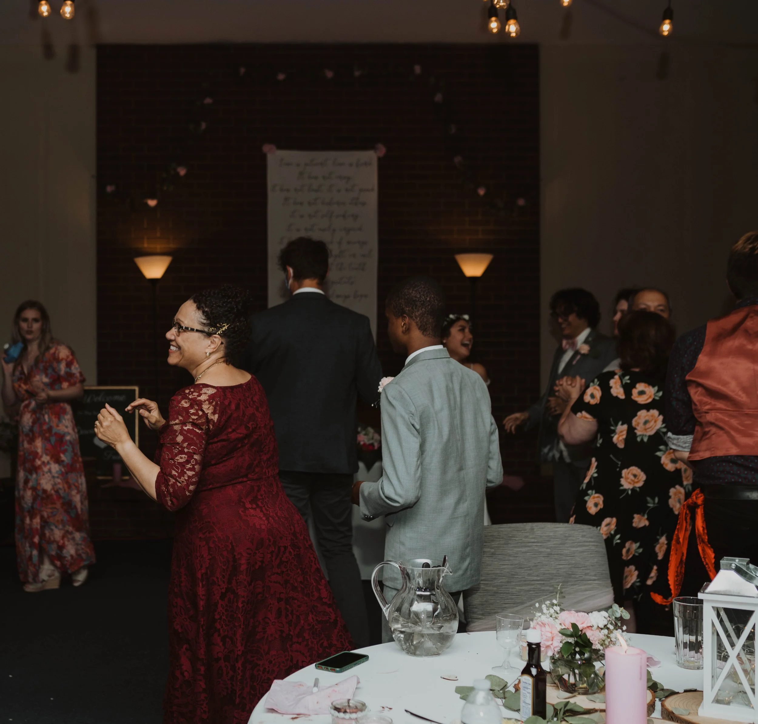 People dancing and socializing at a wedding reception or celebration in a dimly lit room, with a decorated table with flowers, candles, and drinks visible in the foreground. Seattle, WA wedding photography.