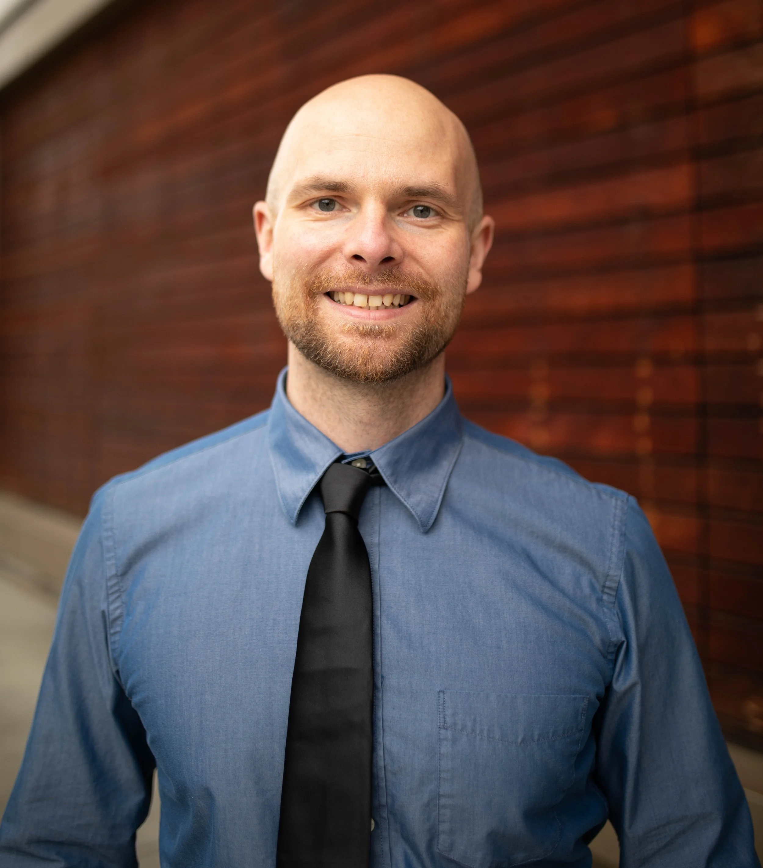 A smiling man with a beard and shaved head wearing a blue dress shirt and a black tie standing outdoors in front of a wooden wall. Seattle professional head shot photography