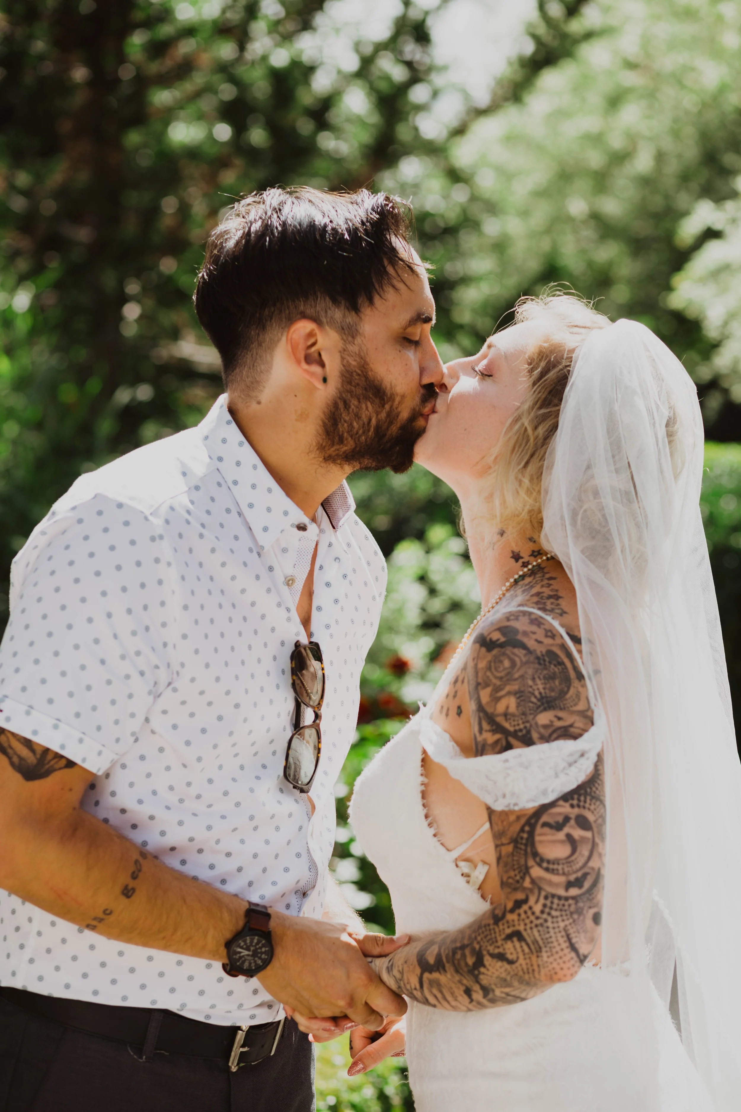 A man and woman sharing a kiss outdoors; the woman is wearing a wedding dress and veil, and has tattoos on her arms; the man is wearing a white polka dot shirt with sunglasses hanging from his collar, a watch, and a bracelet. Seattle, WA wedding phot