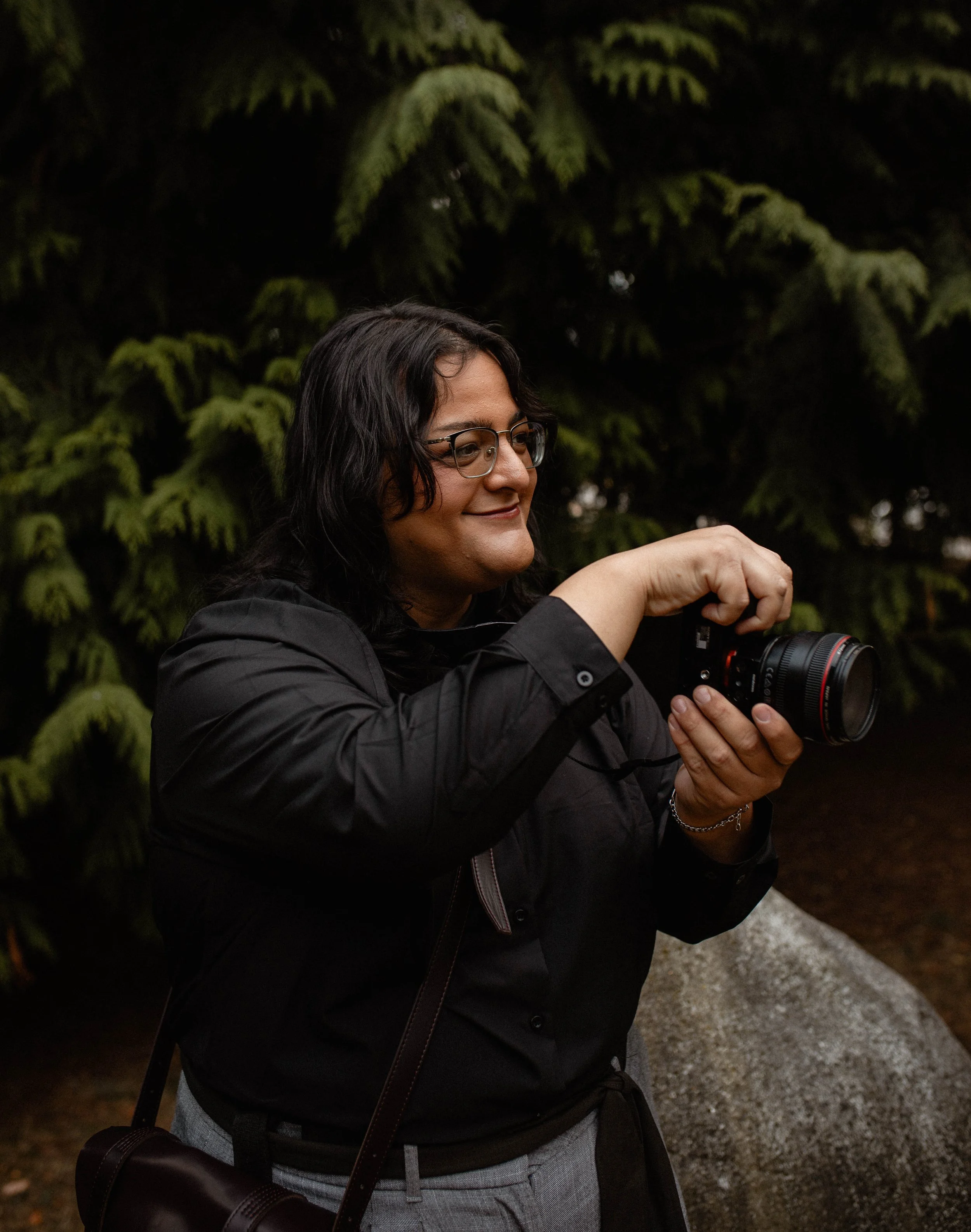 A woman with dark hair, glasses, and a black jacket takes a photograph with a camera outdoors, against a background of green foliage. Seattle professional head shot photography