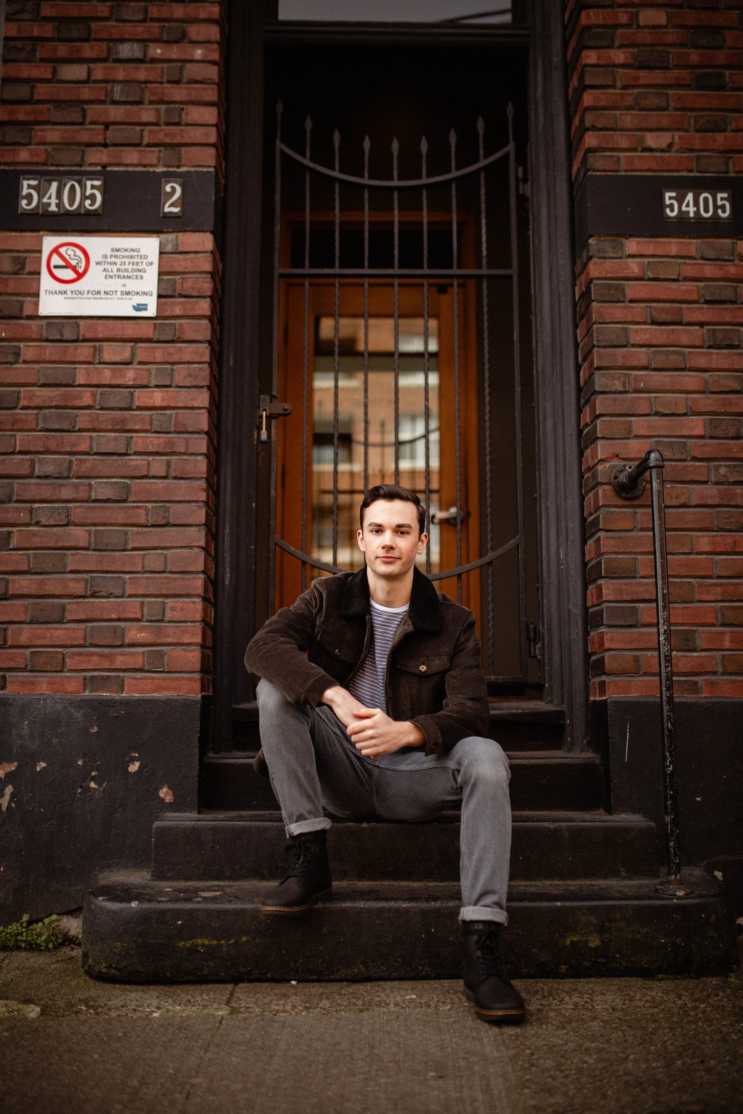 A young man with dark hair, wearing a dark jacket, striped shirt, gray jeans, and black boots, sitting on the steps of a brick building with a black metal gate and distinctive house numbers 5405 and 2. Seattle professional head shot photography.