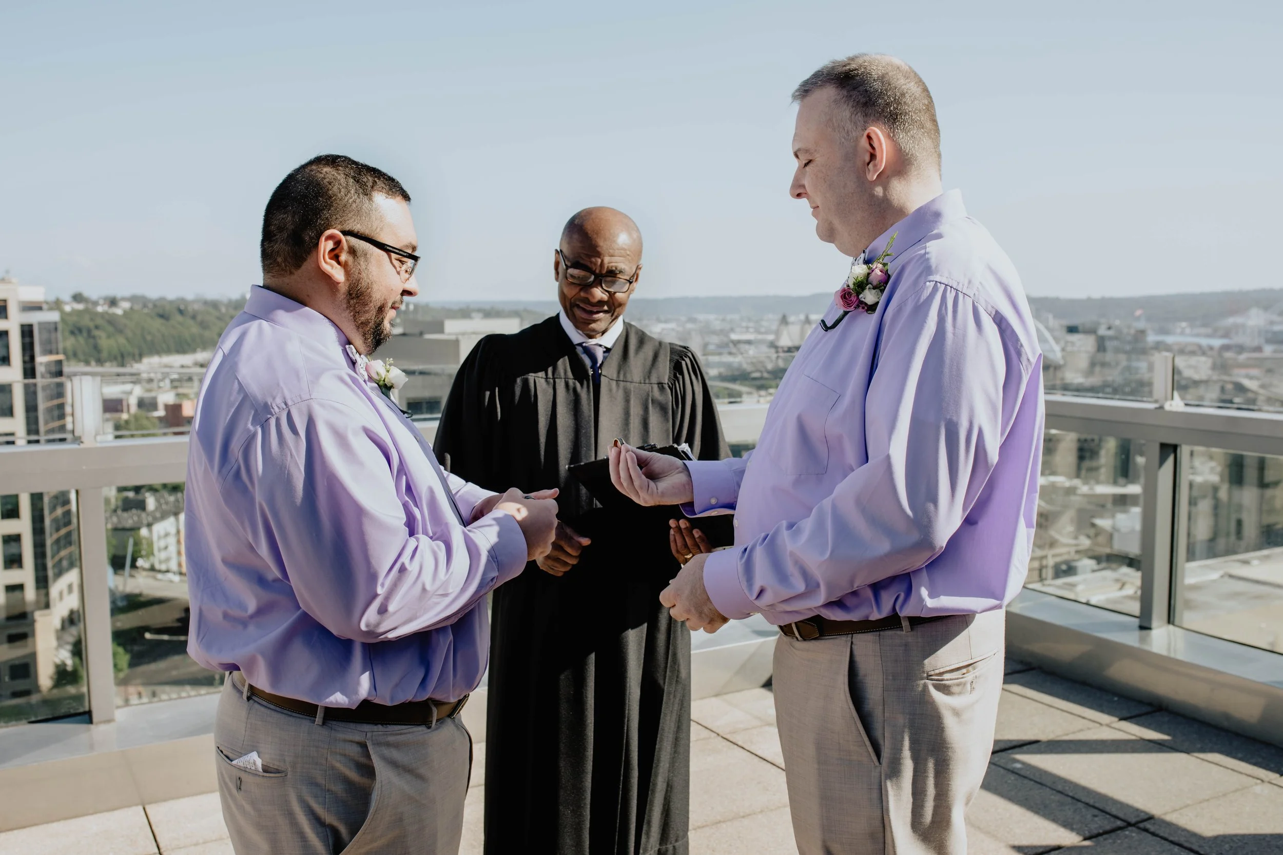 Two men in lavender dress shirts and beige pants exchange rings during a wedding ceremony on a rooftop, with a officiant and city skyline in the background. Seattle Municipal Courthouse wedding photography.