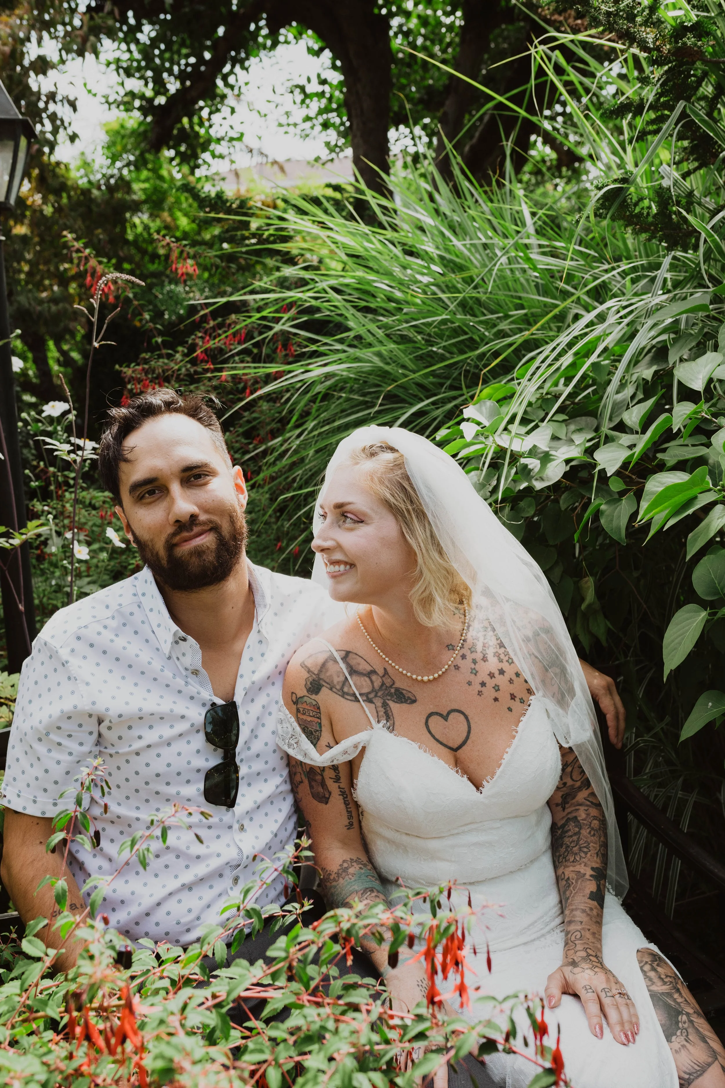 A man and a woman in wedding attire sitting outdoors among green plants, with the woman looking at the man and smiling. Seattle, WA wedding photography.