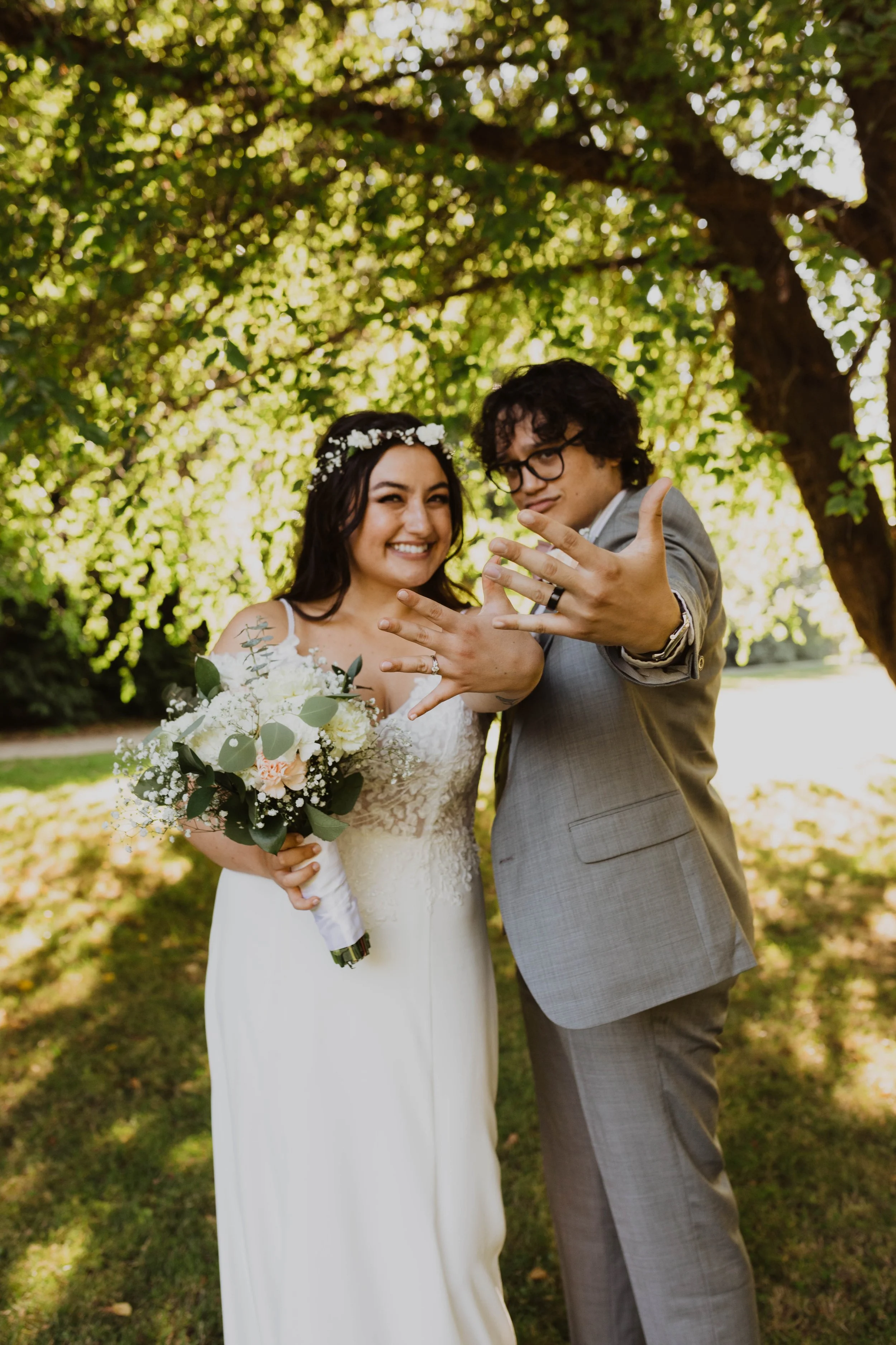 A newlywed couple showing off their wedding rings outdoors under a leafy tree. The bride wears a white wedding dress and holds a bouquet of white and green flowers. The groom wears a gray suit and glasses. Both are smiling and celebrating their weddi