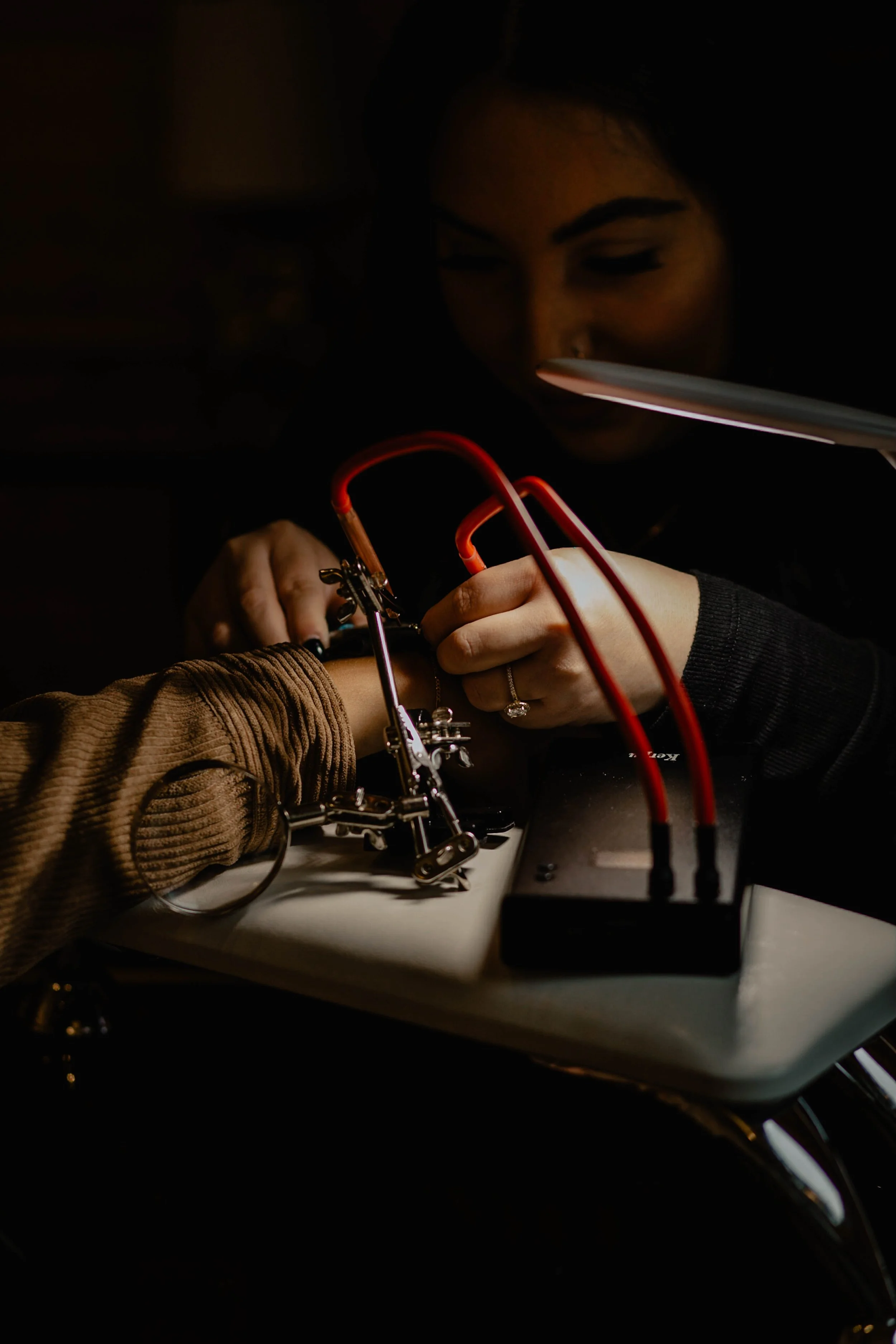 A woman working on sewing or jewelry creation, using a soldering or jewelry-making tool with a third person's arm in the frame, on a white table with electrical wires connected to a power supply. Seattle professional head shot photography