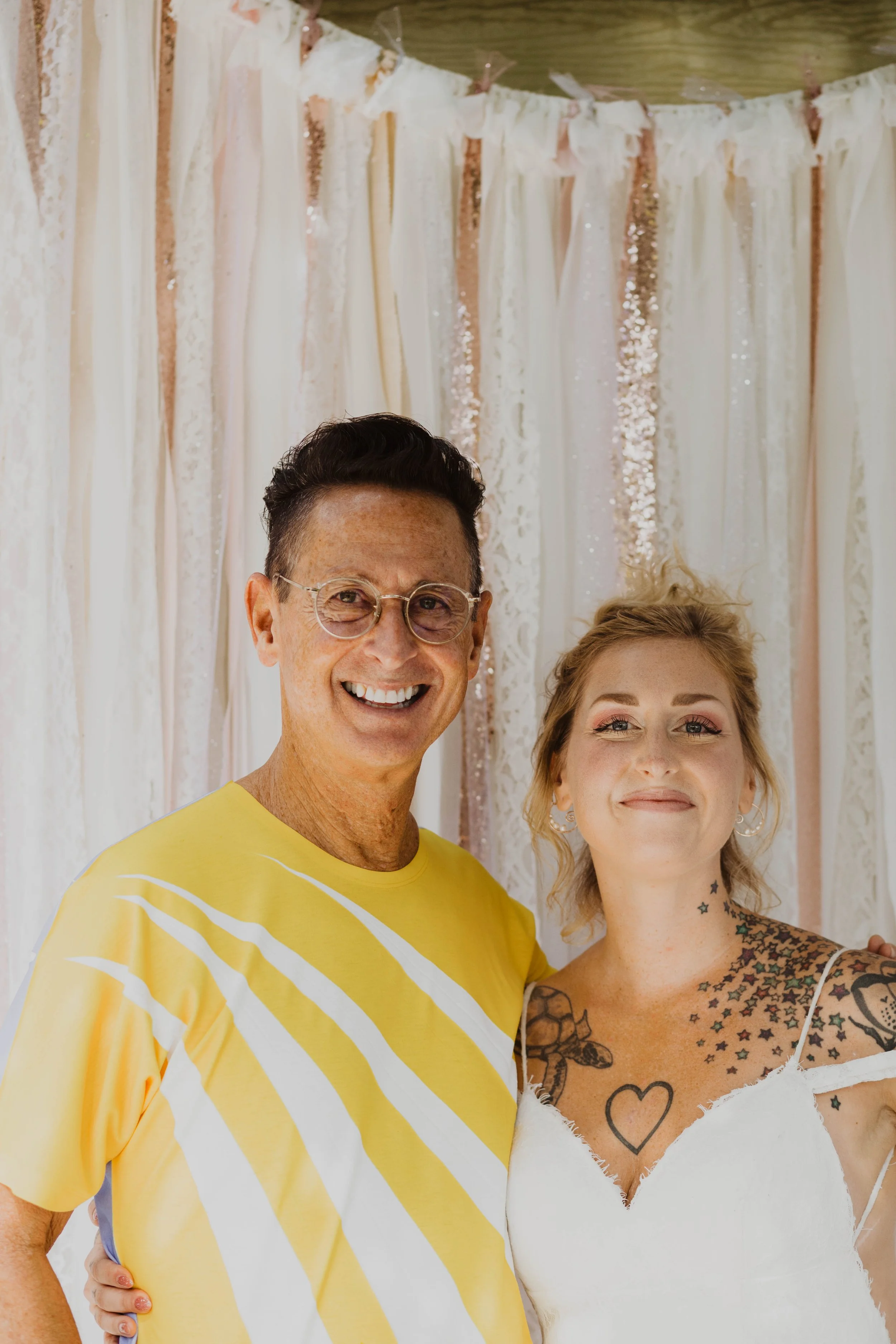 A smiling man and woman standing in front of a decorative backdrop of cream and rose gold streamers, with the woman wearing a white dress and visible tattoos. Seattle, WA wedding photography.