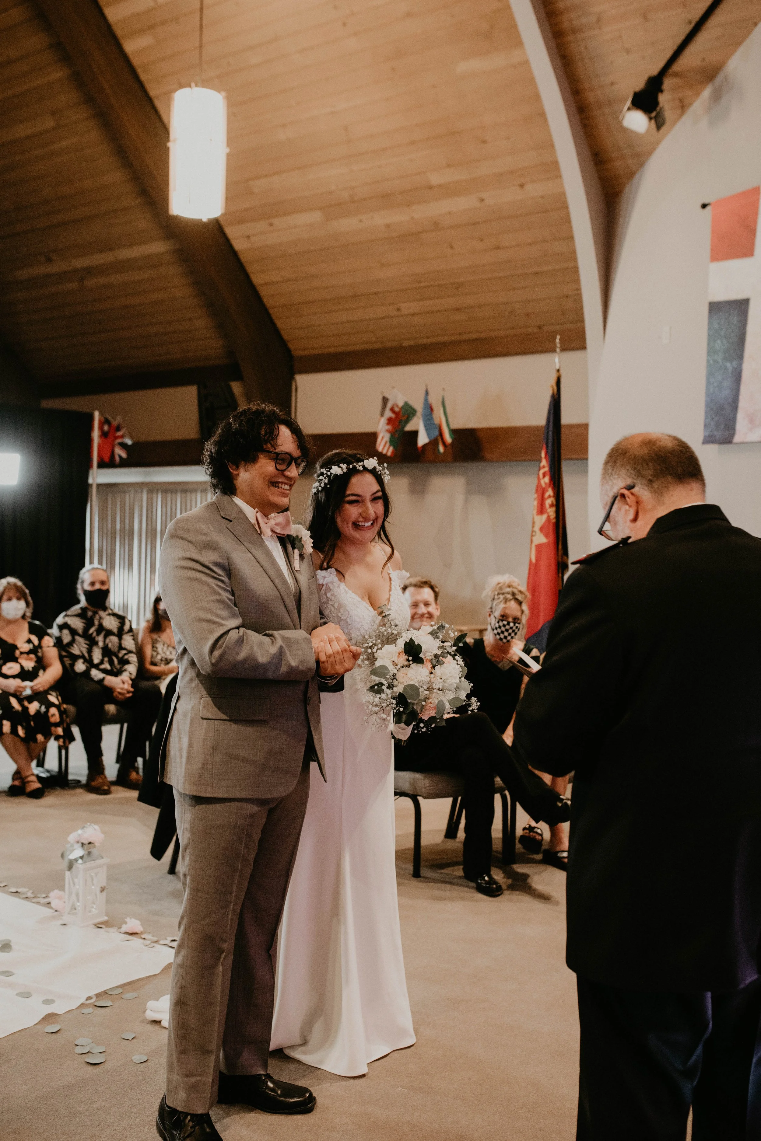A couple getting married, smiling, during a ceremony in a church or hall with guests seated in the background, some wearing masks. The bride wears a white dress with a floral headband and holds a bouquet, while the groom wears a light-colored suit wi