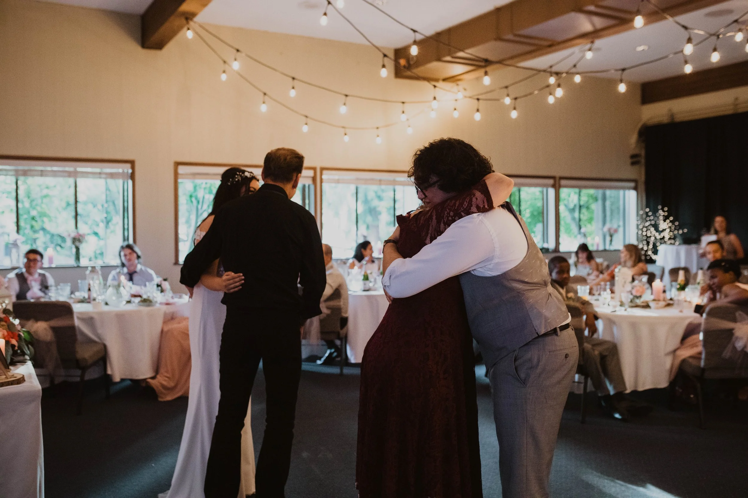 People hugging at a wedding reception inside a decorated hall with string lights and seated guests Seattle, WA wedding photography.