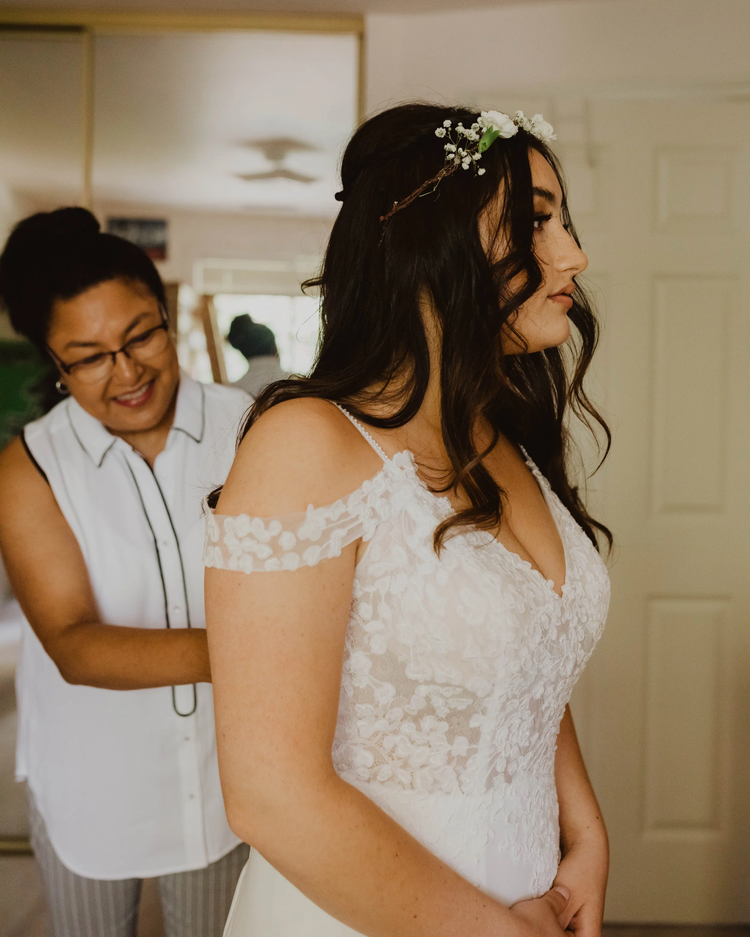 A woman in a white lace off-shoulder dress with a floral headpiece is having her dress adjusted by another woman in glasses, wearing a sleeveless white shirt, in a room with beige walls. Seattle, WA wedding photography.