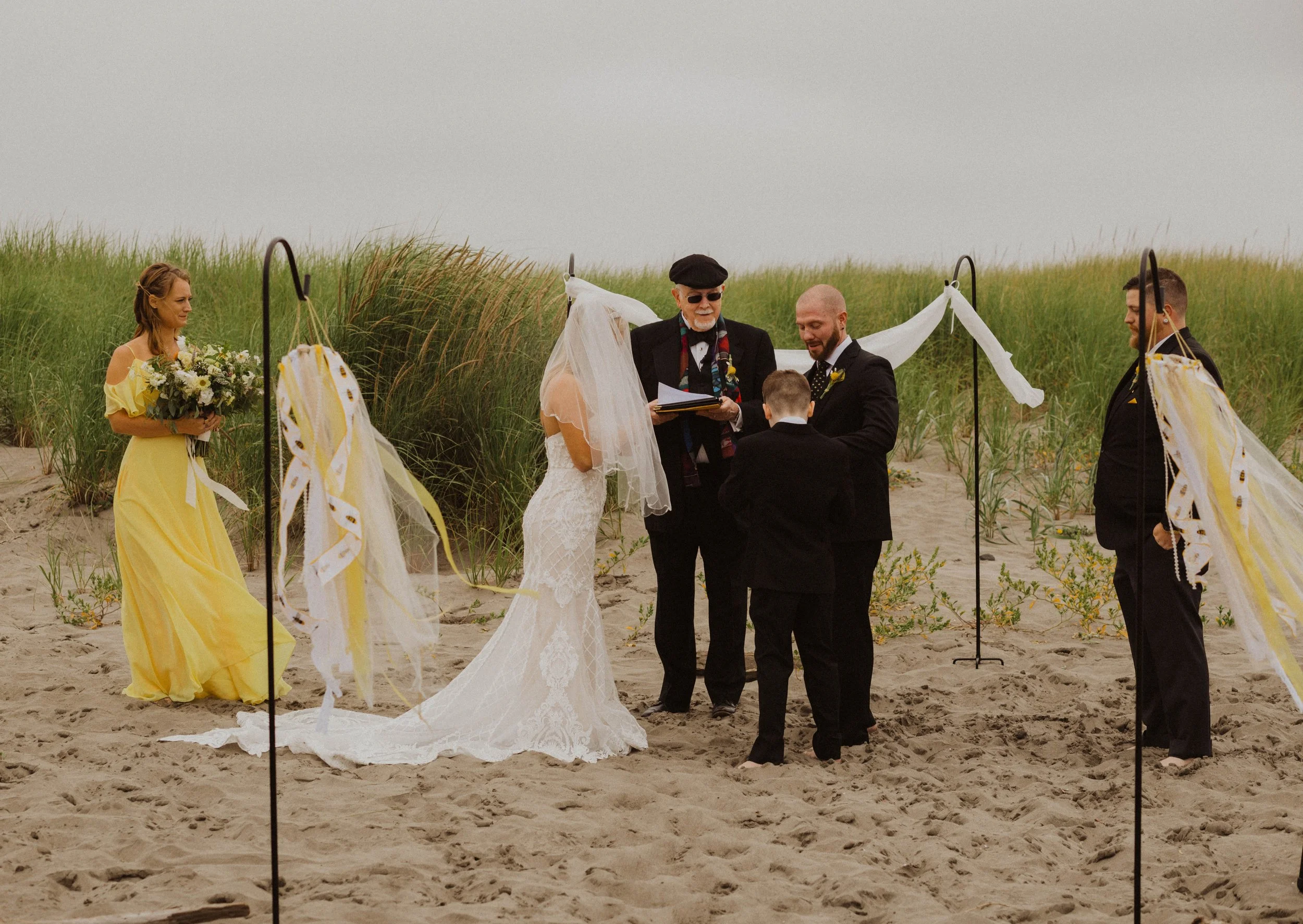 A beach wedding ceremony with a bride and groom standing beneath a simple arch, officiant reading from a book, bridesmaid holding a bouquet, and groomsmen standing on the sand with grassy dunes in the background. Long Beach, WA wedding photography.