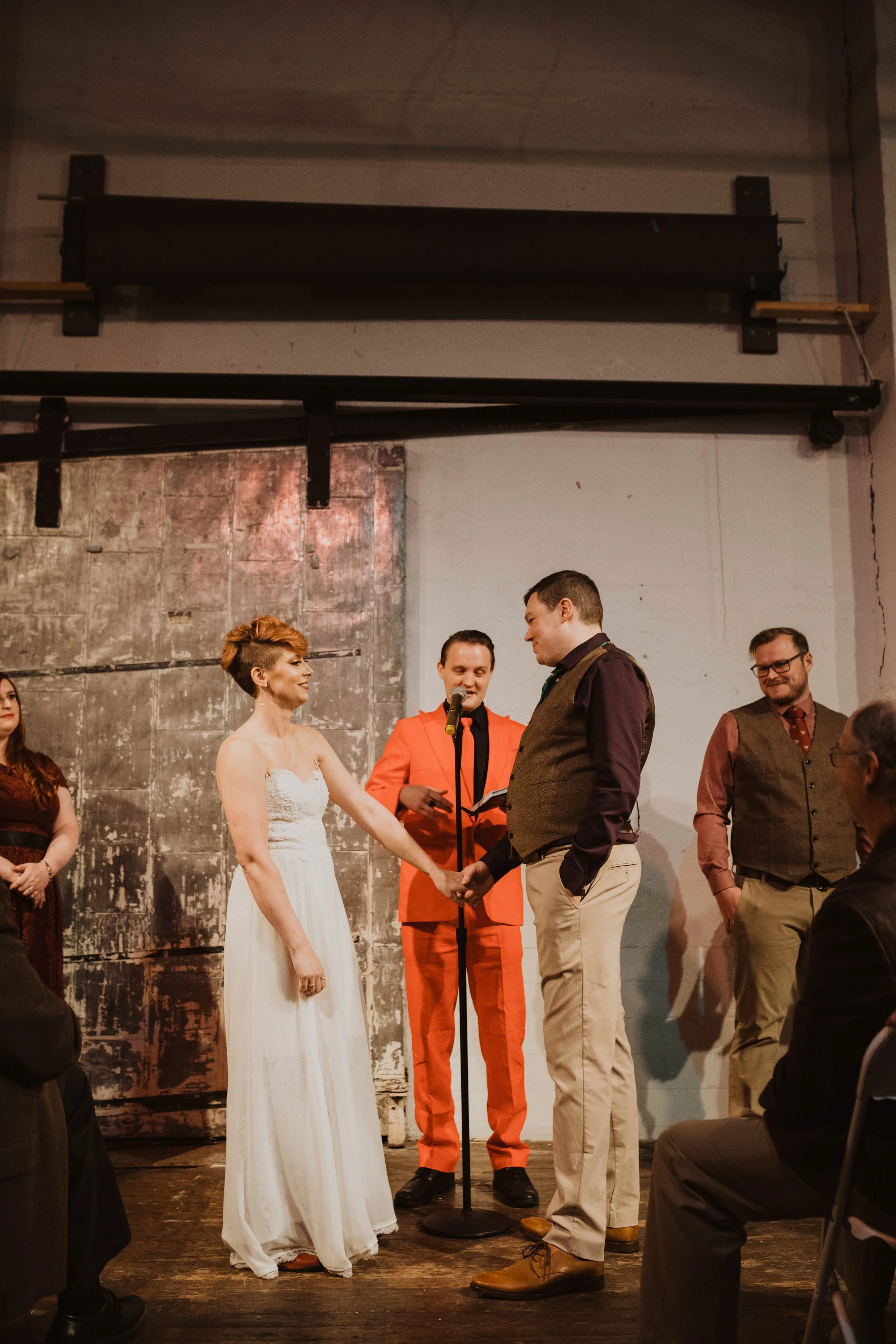 A couple holding hands during their wedding ceremony, with an officiant reading from a book, in a rustic indoor setting with other guests and wedding party members present. Pioneer Square, Seattle, WA wedding photography.