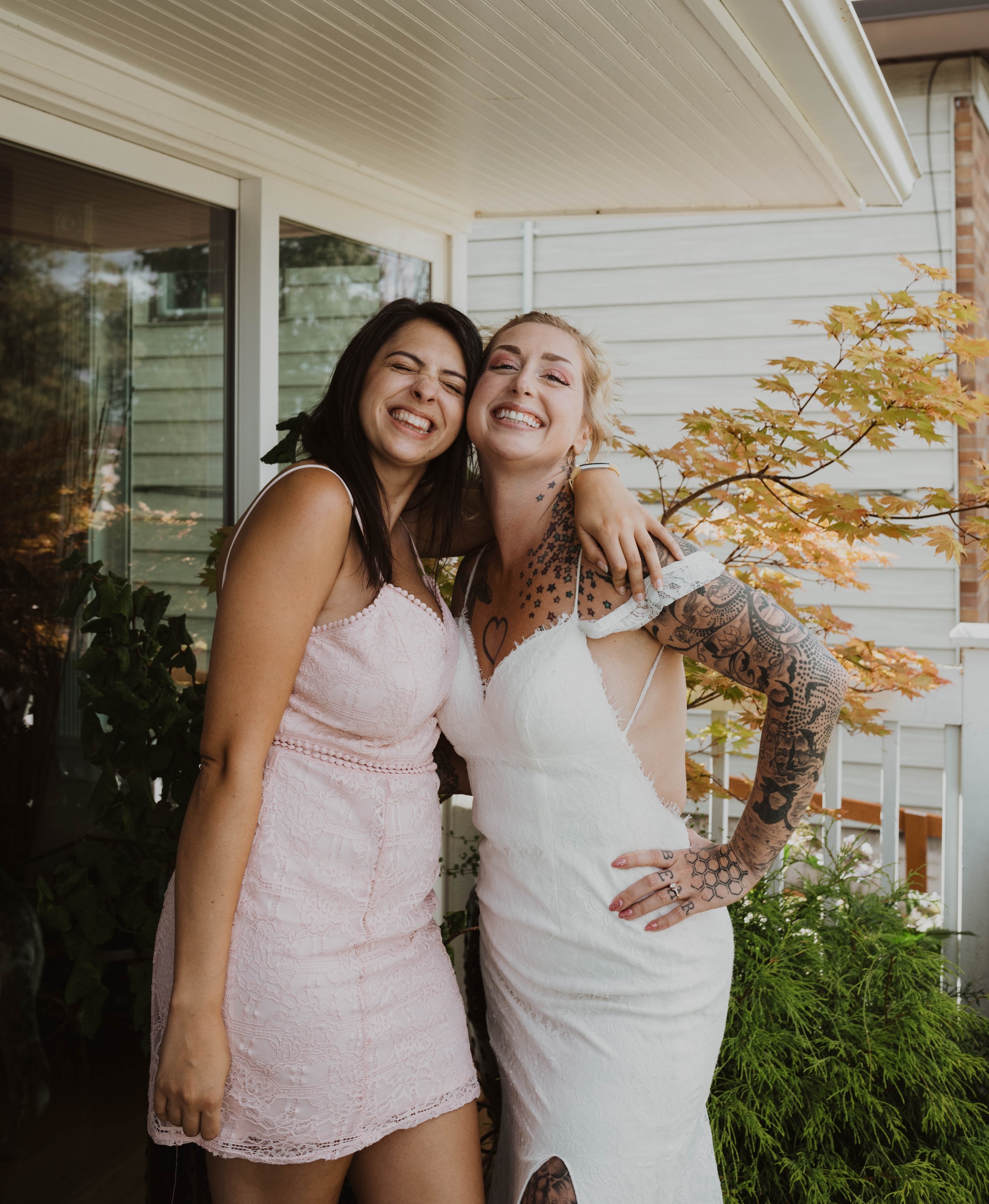 Two women smiling and hugging outdoors in front of a house, one in a pink dress and the other in a white dress with tattoos on her arms. Seattle, WA wedding photography.