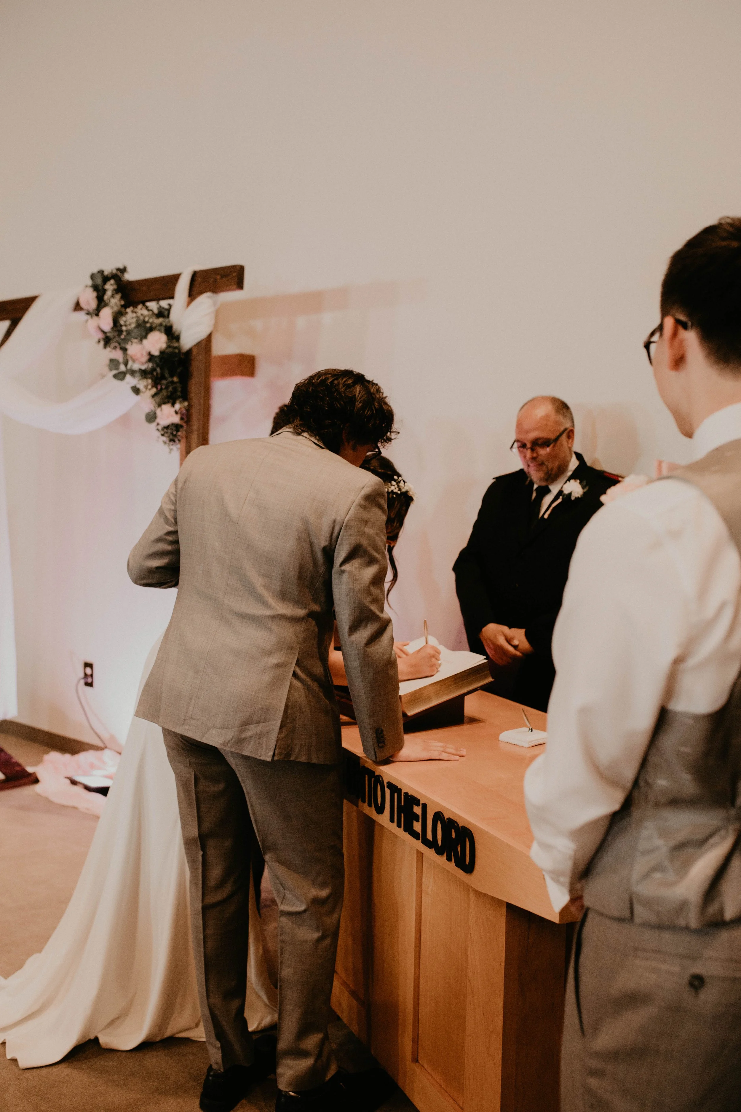 A wedding ceremony with a couple signing a marriage register at a wooden desk labeled 'NOTARY PUBLIC' while an officiant and guests watch, decorated with flowers and fabric in a hall. Seattle, WA wedding photography.