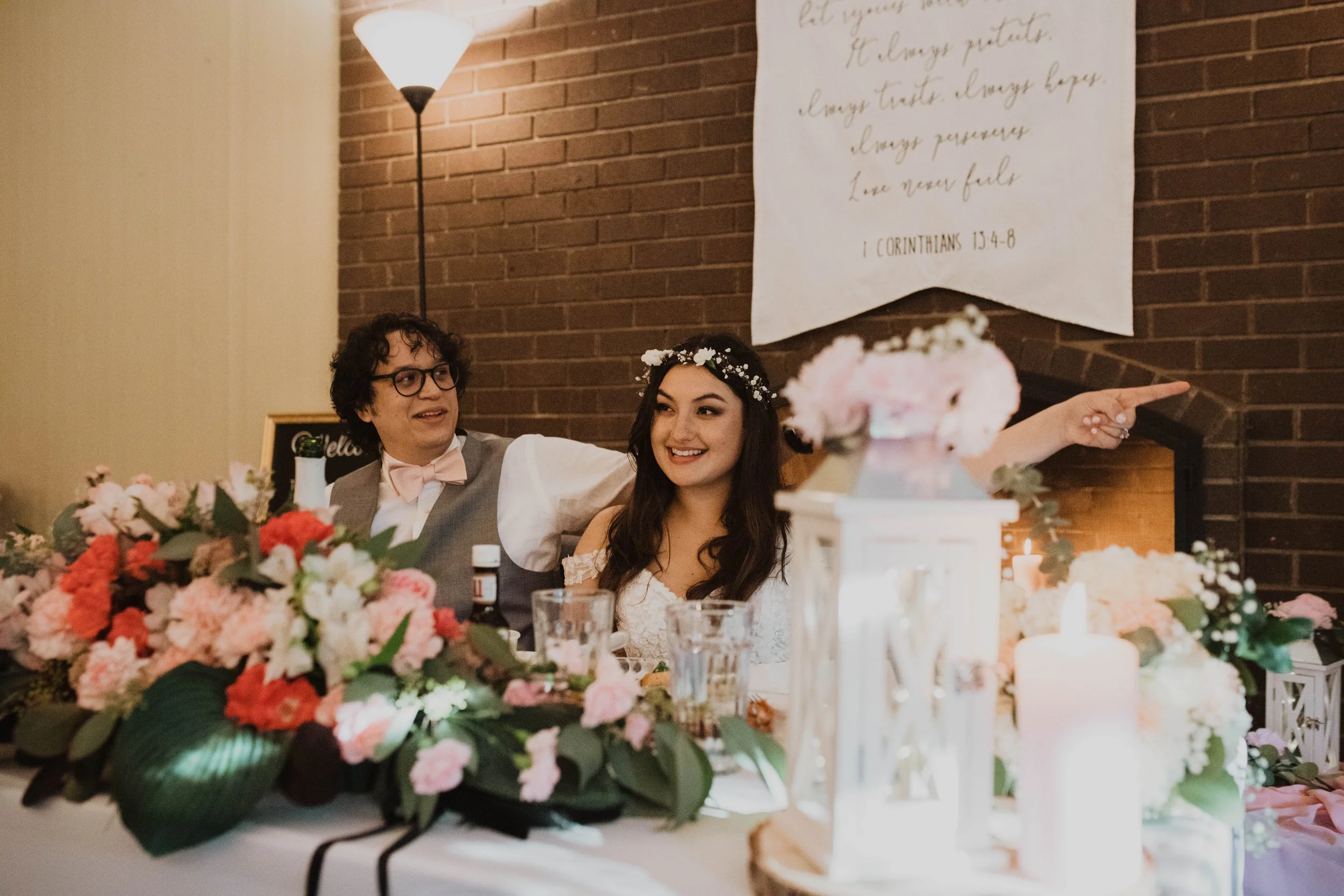 A smiling woman wearing a flower crown sits at a decorated wedding table with a man pointing to the right. The table has pink and white flowers, candles, and a lantern. A brick wall with a hanging banner features a Bible verse in the background.