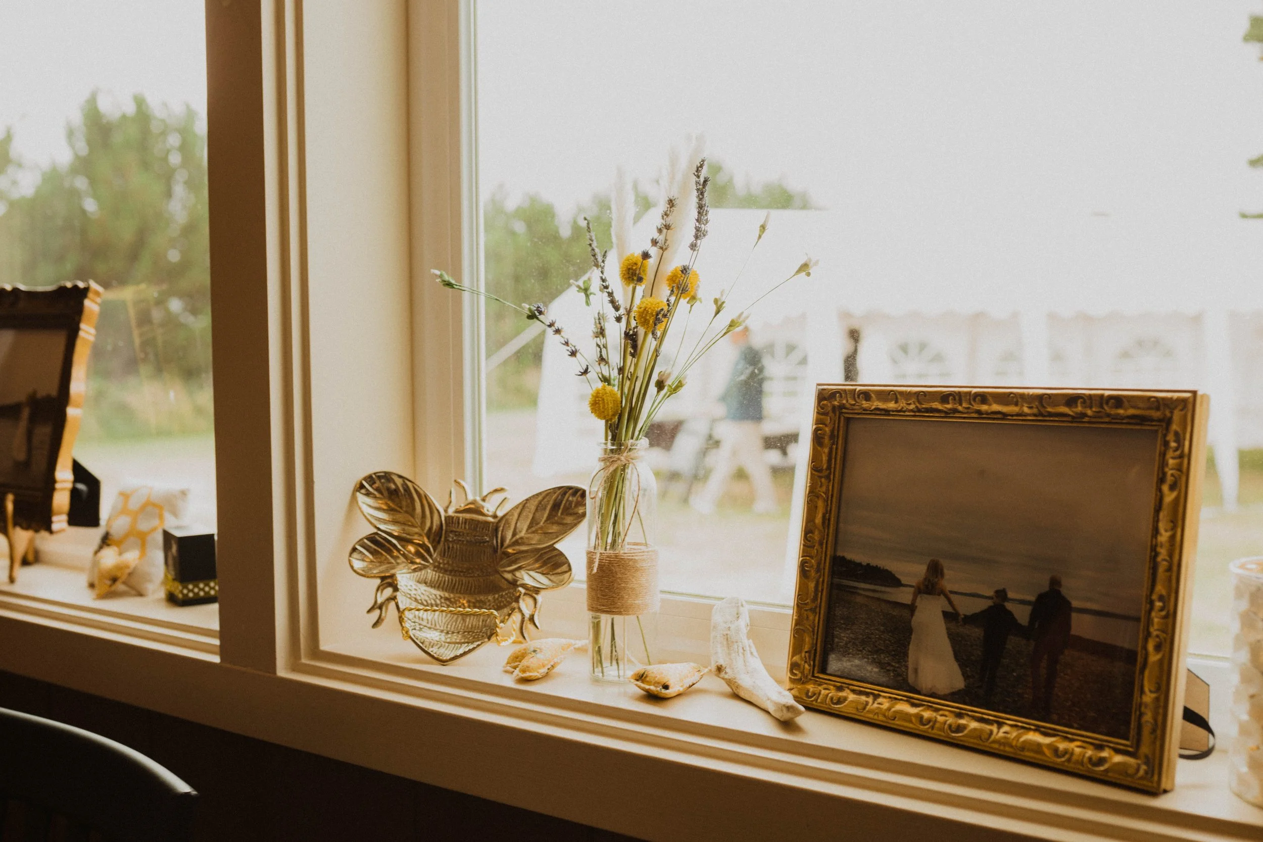 Decorative items on a window sill: a glass jar with dried flowers, a gold butterfly-shaped dish, a framed photograph of three people on a beach, seashells, and a coral piece; outside the window, there is a grassy area and a white tent or structure.