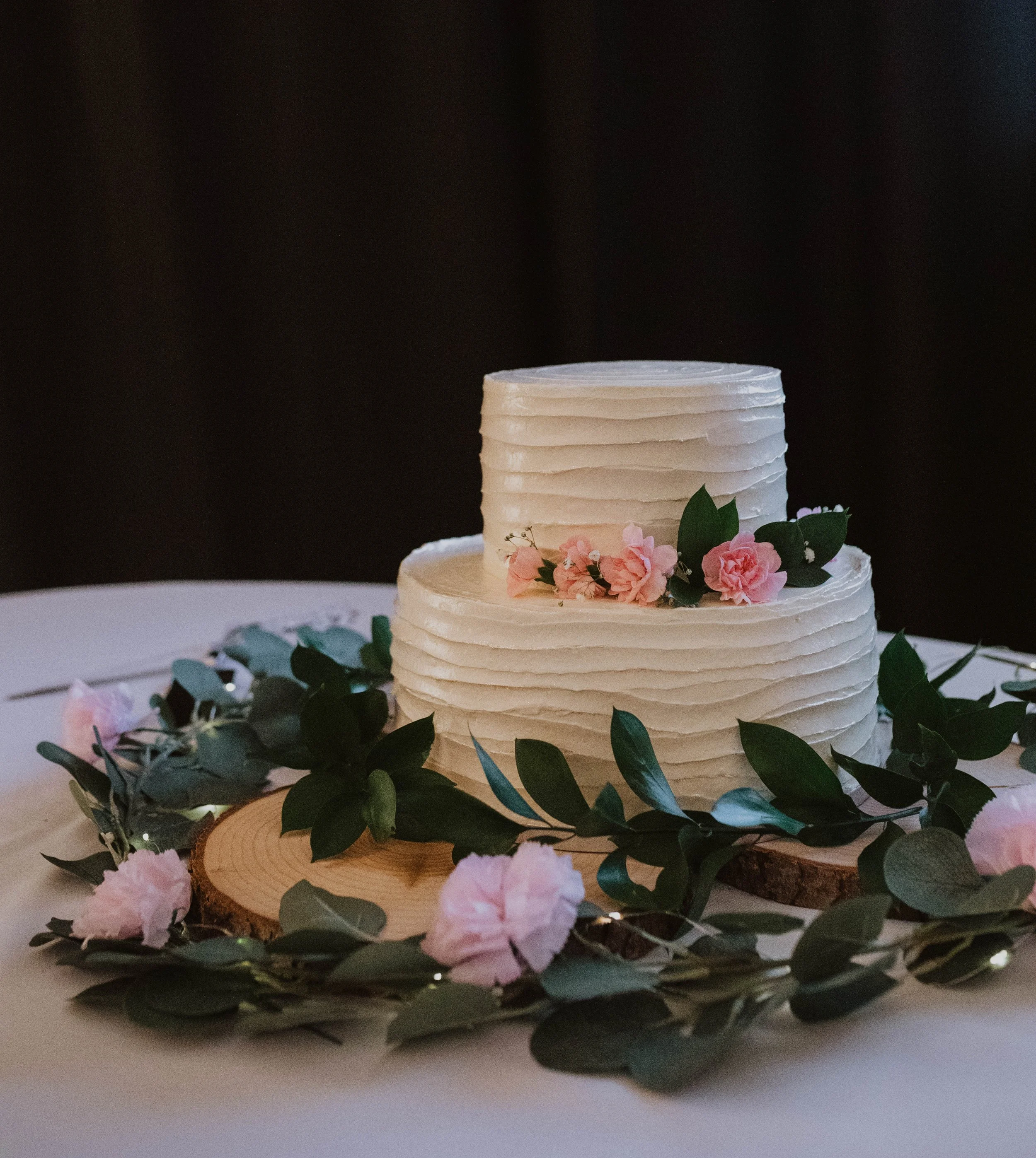 Two-tiered white wedding cake with textured frosting, decorated with pink flowers and green leaves, on a wooden slab surrounded by greenery and pink flowers. Seattle, WA wedding photography.