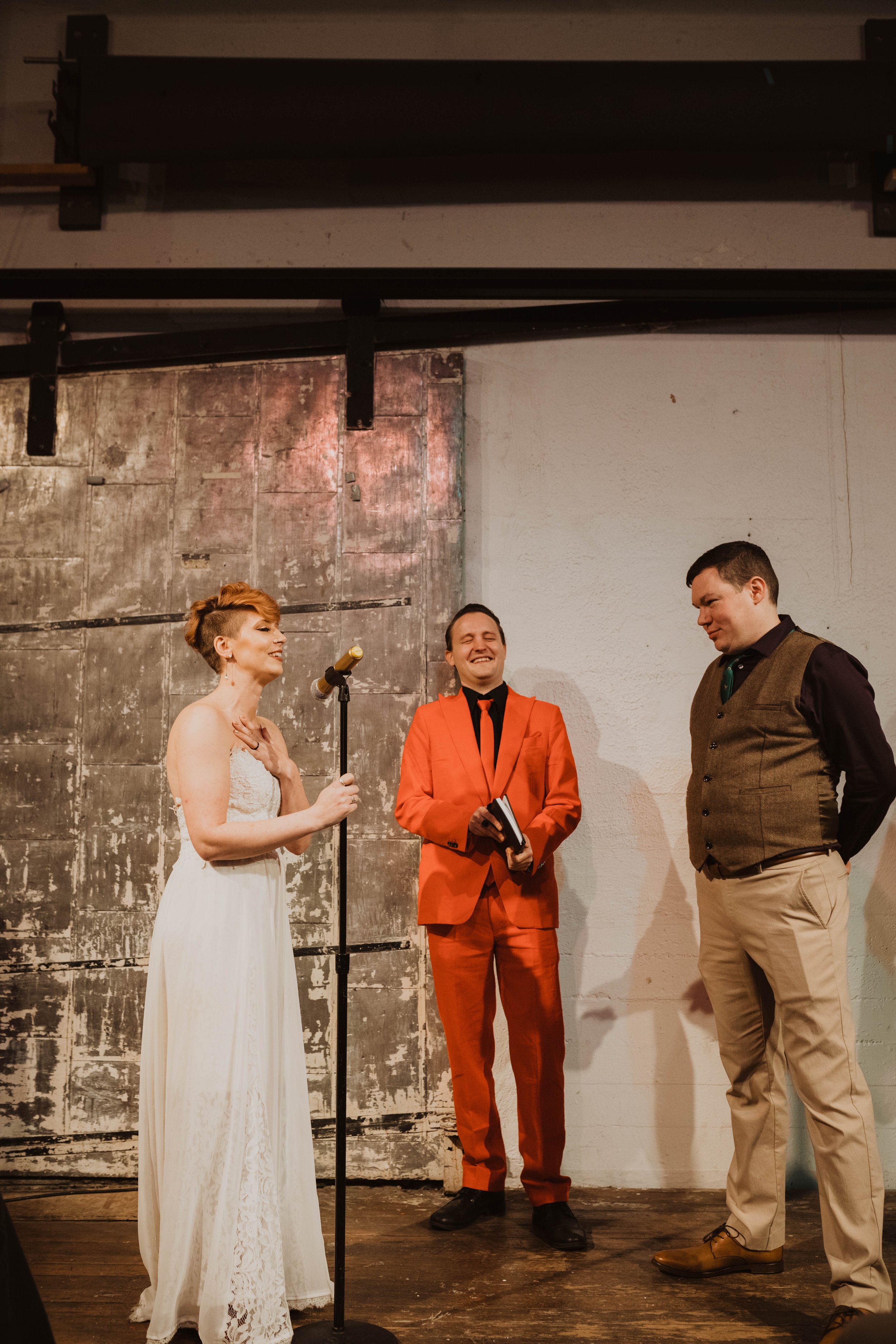 A woman in a white dress and two men, one in a red suit and the other in a brown vest and beige pants, standing on a stage in front of a rustic backdrop, with the woman holding a microphone and laughing.