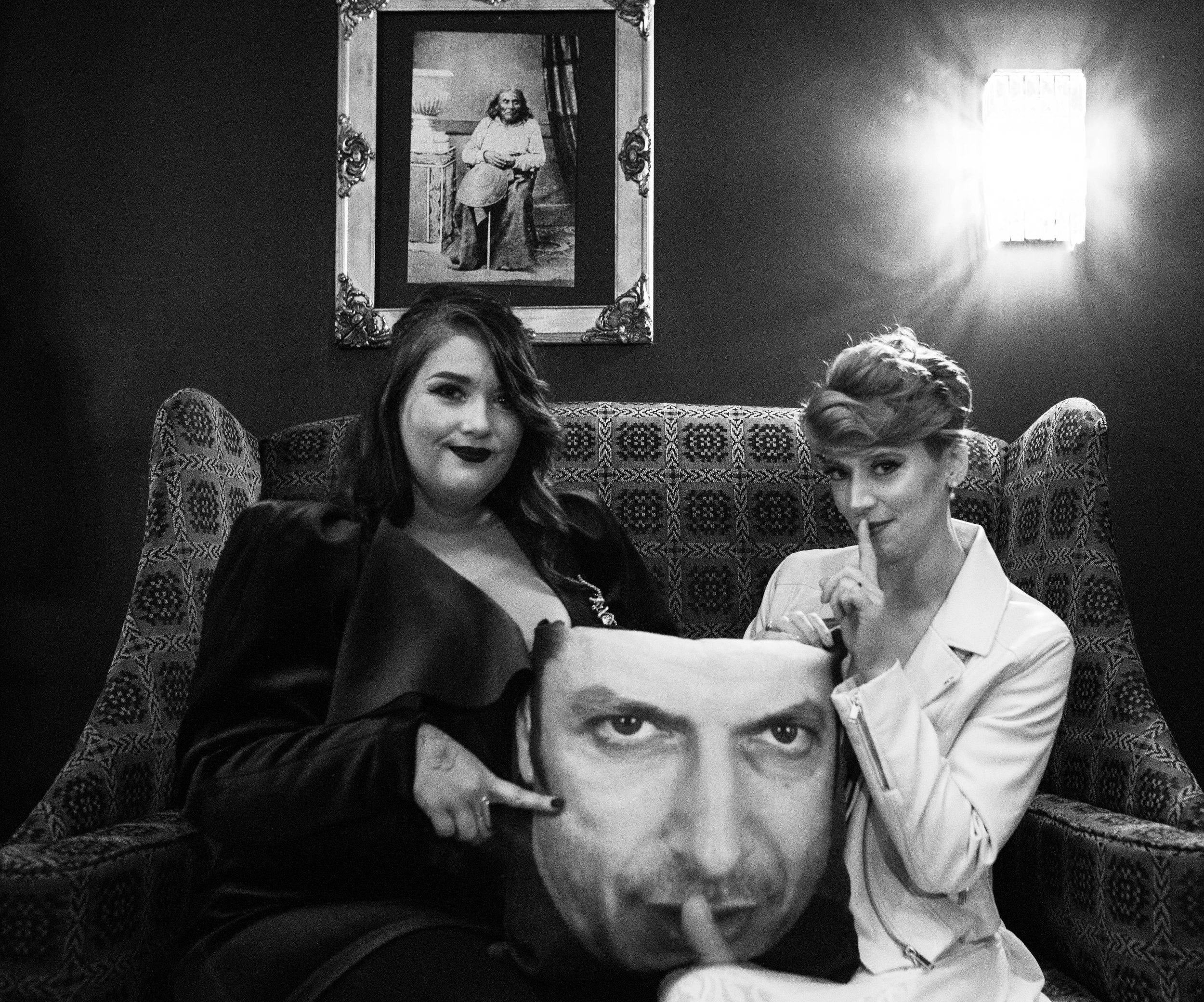 Two women sitting on a patterned couch in a dimly lit room. One woman is holding a pillow with a large faceshot print, and the other woman is holding a pillow with a person's face. The woman on the right has her index finger to her lips. There is a f