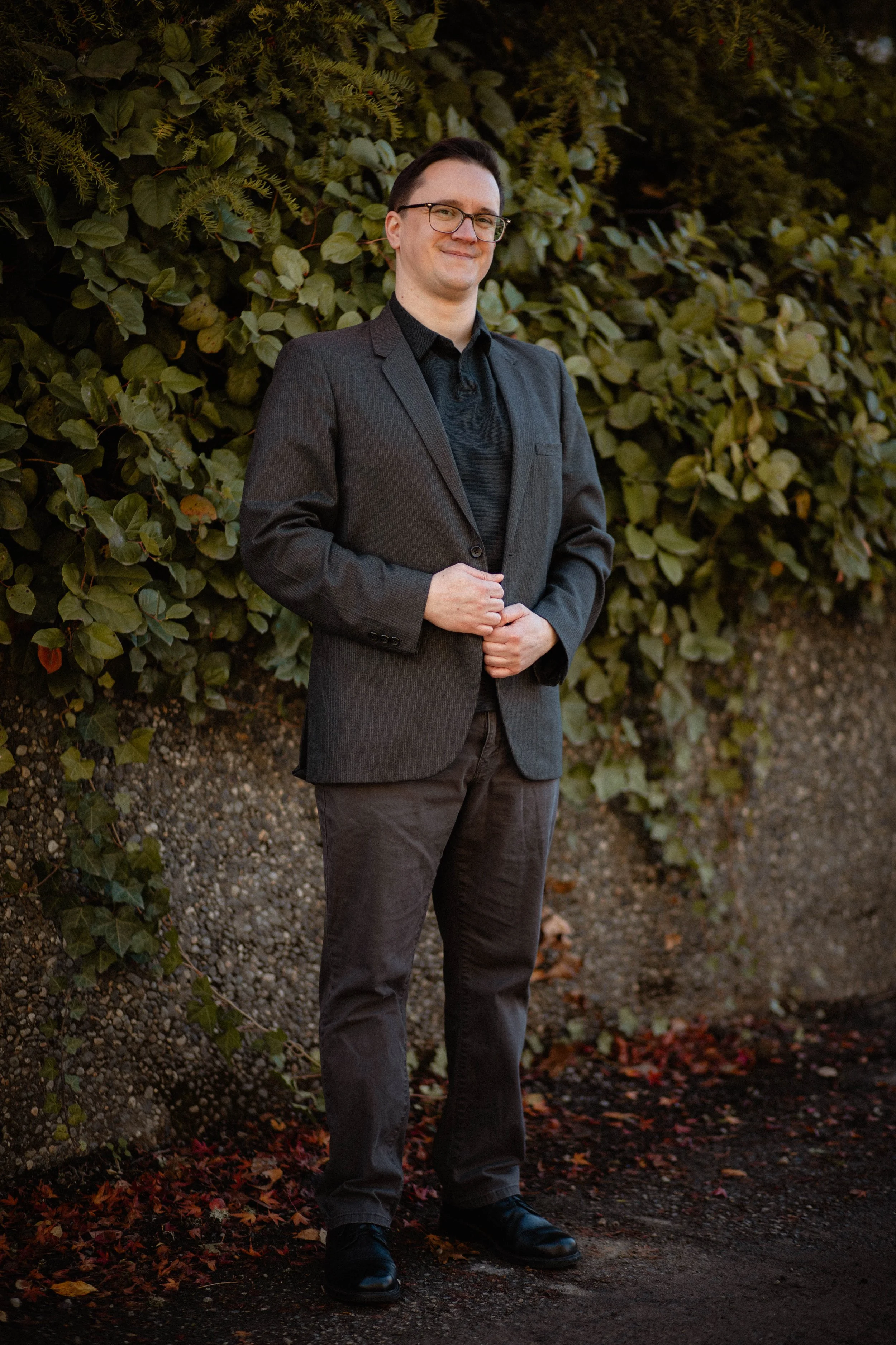 A man in a dark suit and glasses standing outdoors in front of a leafy hedge, smiling with his hands loosely clasped. Seattle professional head shot photography