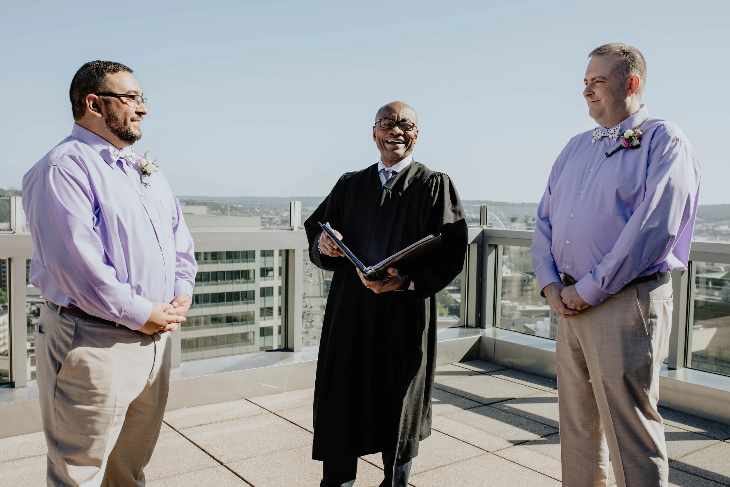 A same-sex wedding ceremony on a rooftop with a male officiant in a black gown and two men in light purple shirts with bow ties and flowers, with a city skyline in the background. Seattle municipal courthouse wedding photography.