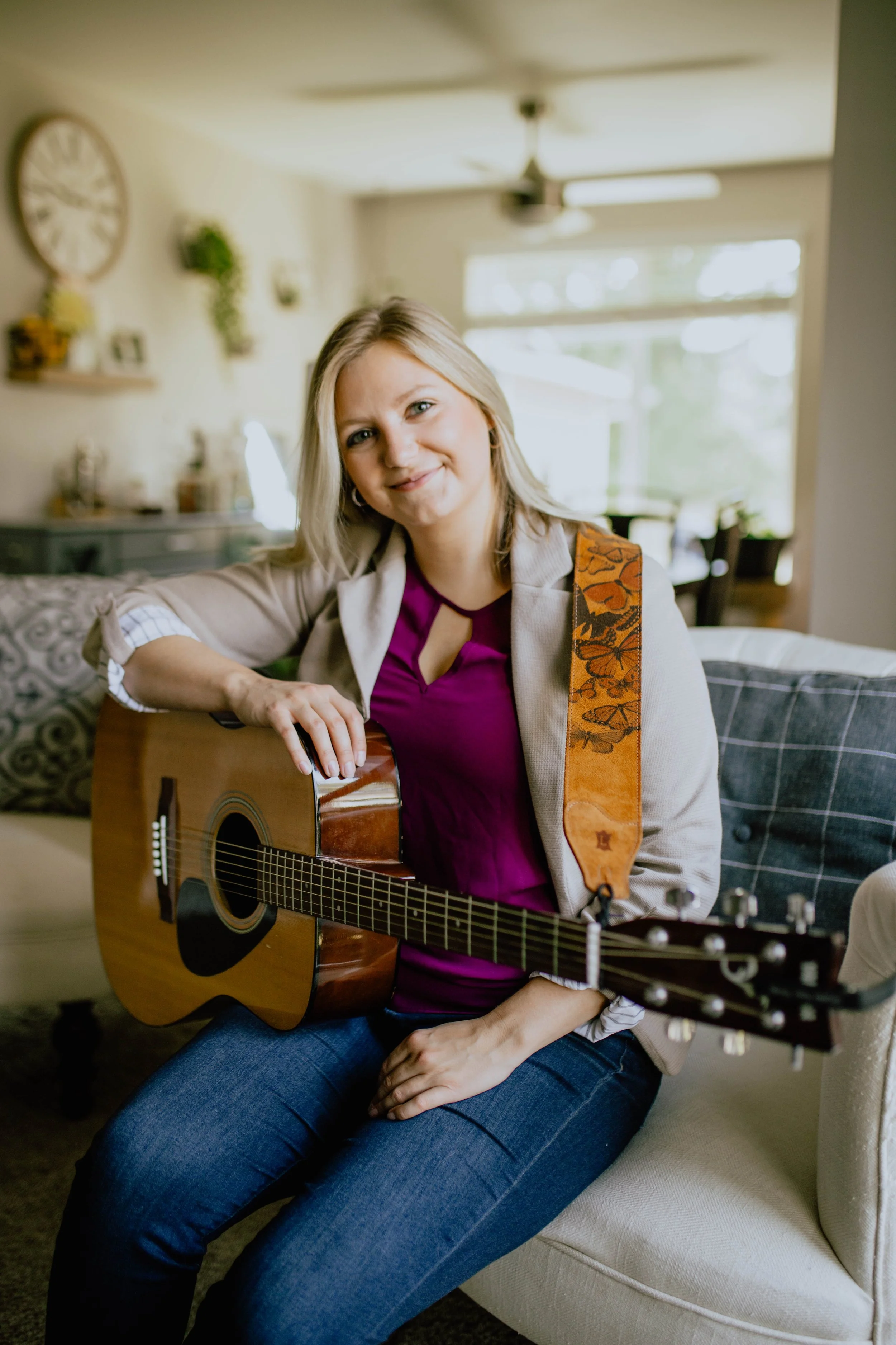 A woman sitting on a sofa holding an acoustic guitar, smiling at the camera in a well-lit, cozy living room. Seattle professional head shot photography