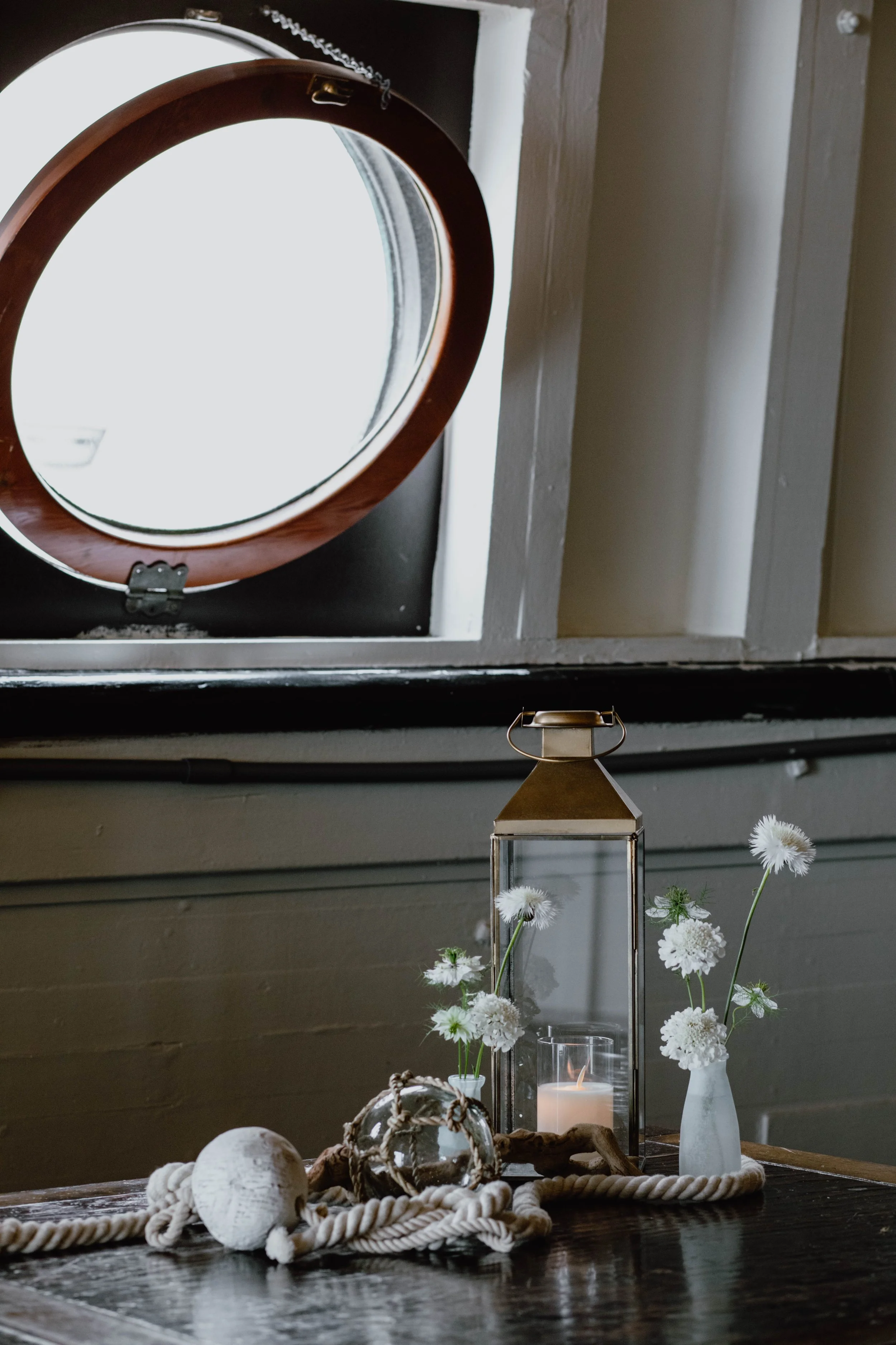 Decorative arrangement with white flowers in vases, a lit candle in a glass holder, a rope, and a glass orb on a wooden table, with a round mirror reflecting a window in the background. Seattle event photography