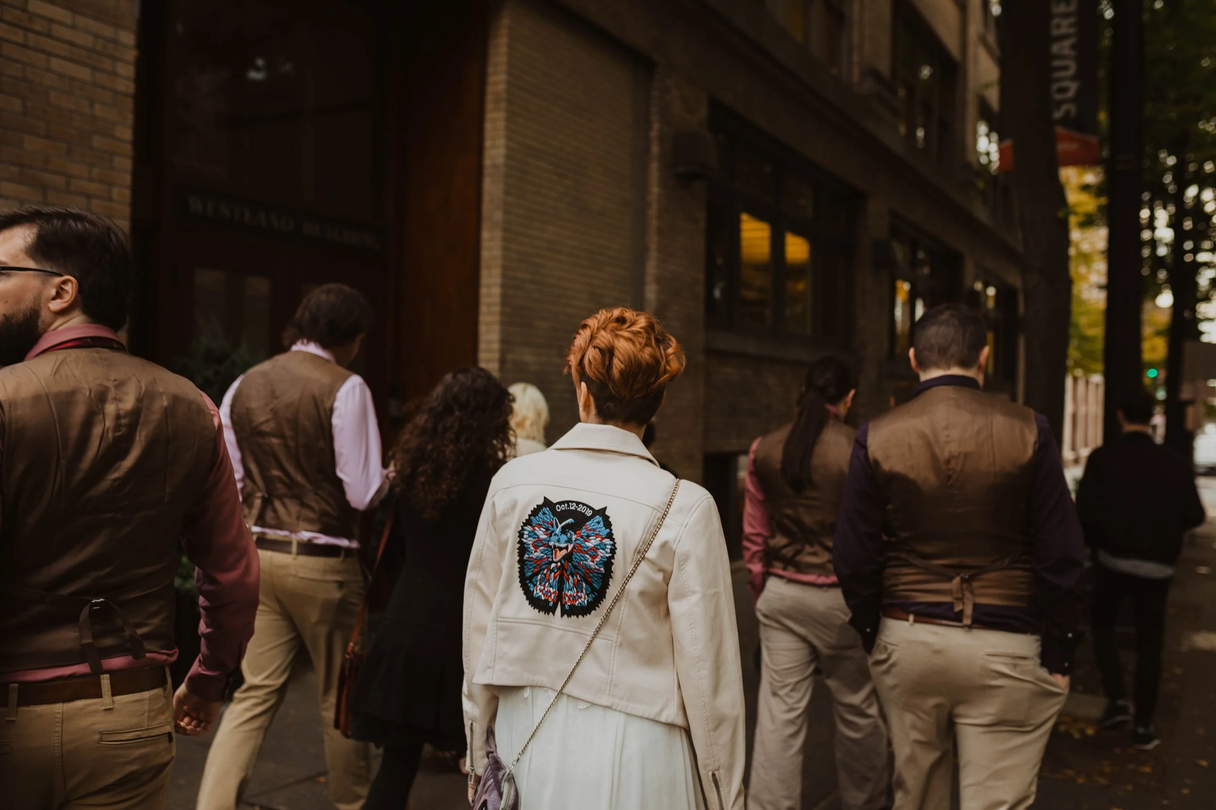 People walking on city sidewalk, seen from behind, with brick building and trees in the background. Pioneer Square, Seattle, WA wedding photography.