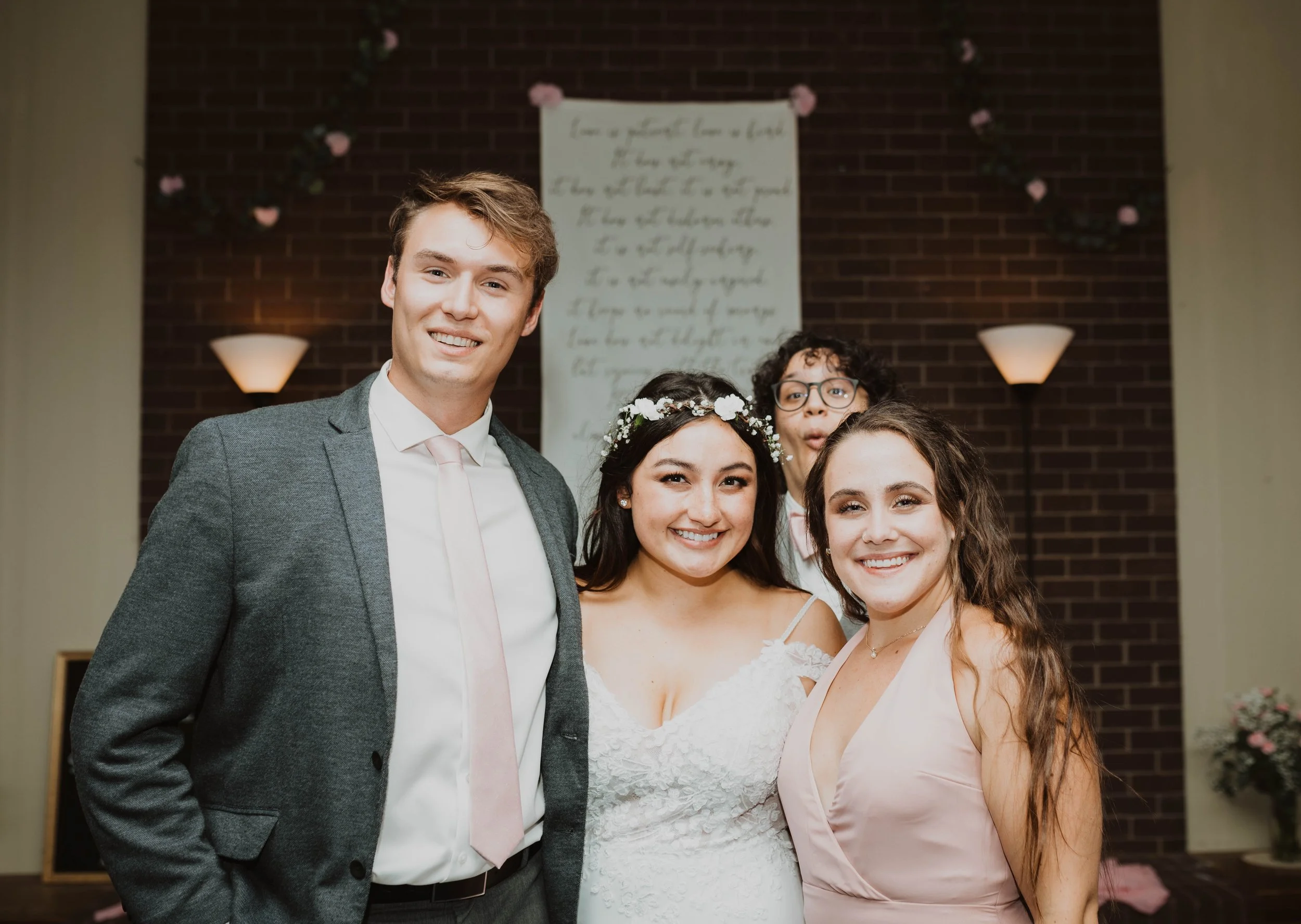 A group of four people smiling at a wedding, with a woman in a white dress and floral headband, and a man in a gray suit, standing together indoors in front of a brick wall with romantic decorations. Seattle, WA wedding photography.