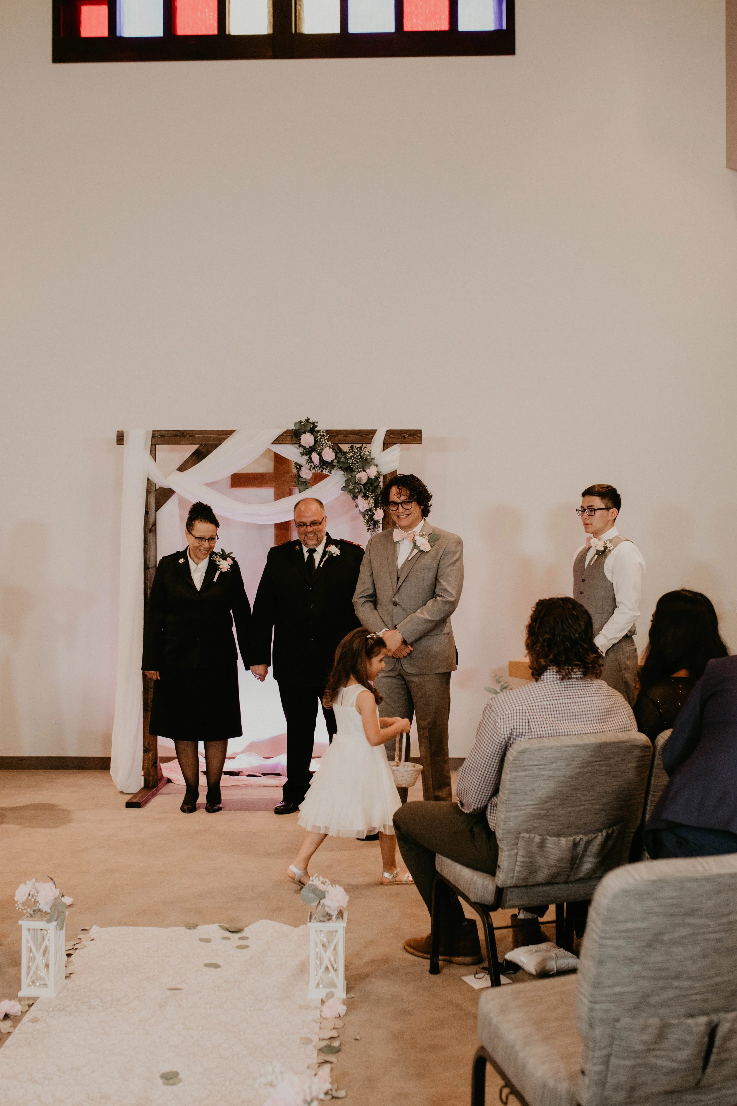 People standing at an indoor wedding ceremony, with a flower arch and cross backdrop, and guests seated in chairs observing. Seattle, WA wedding photography.