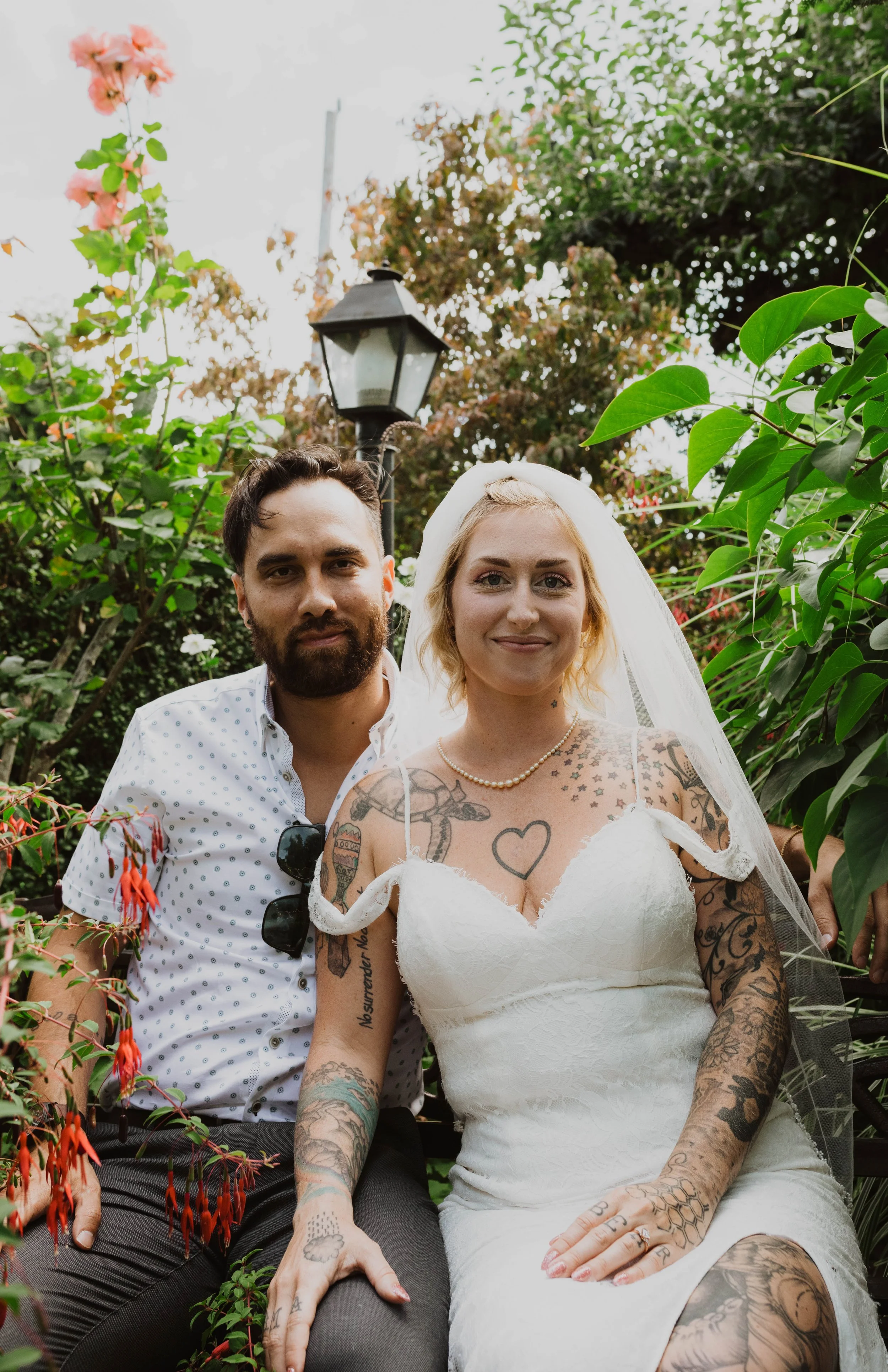 A happy couple, a man with dark hair and beard, and a woman with blonde hair and tattoos wearing a white wedding dress, sitting on a bench in a garden with green foliage and a street lamp in the background. Seattle, WA wedding photography.
