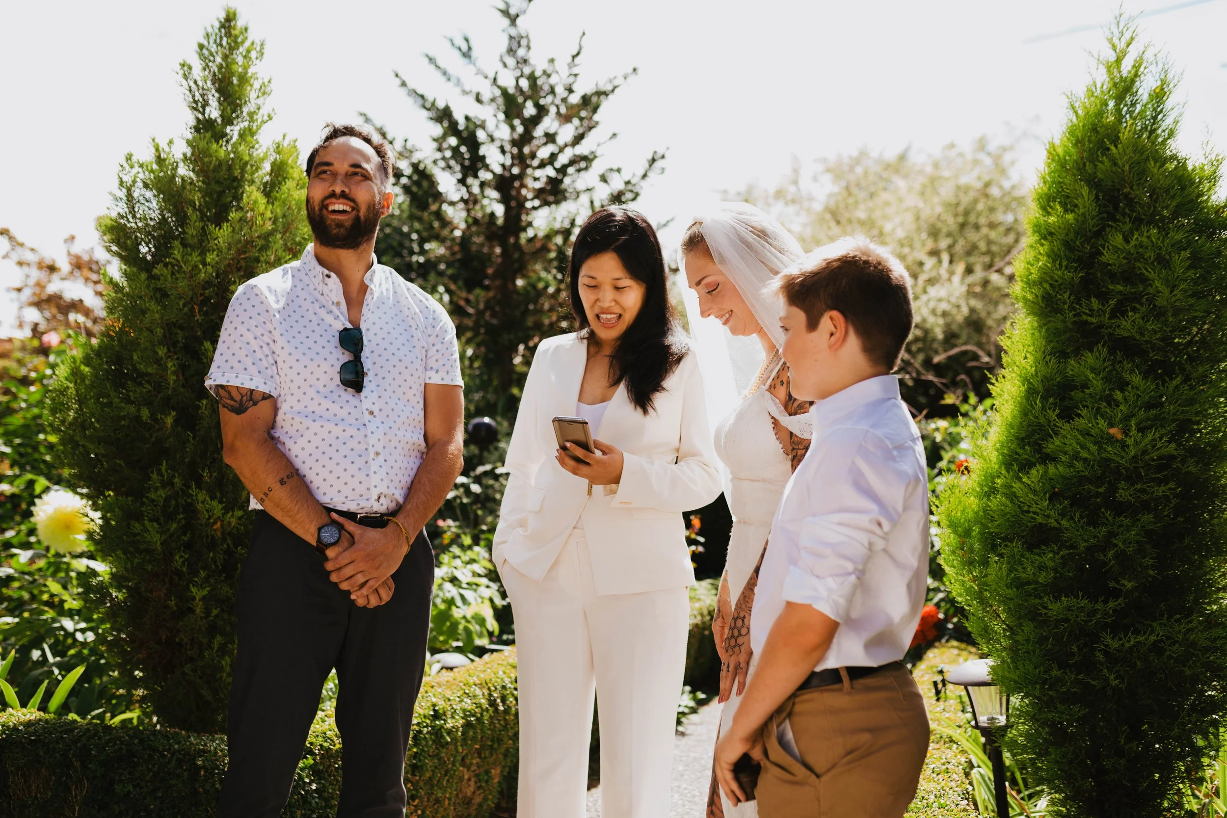 Group of five people standing outdoors, with three women and two men, dressed in summer clothes, looking at a smartphone, surrounded by greenery and trees, on a sunny day.