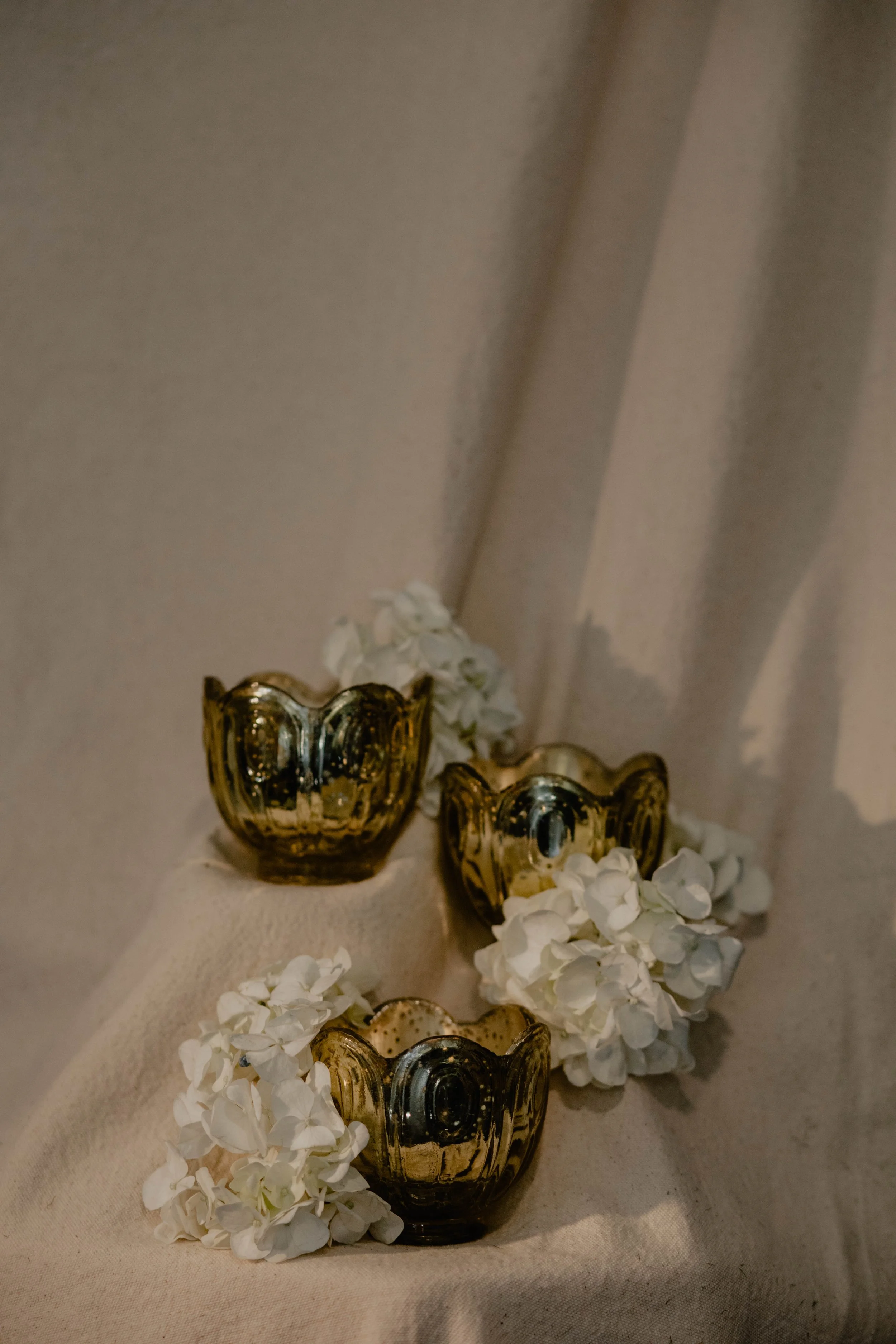 Three gold-colored glass candle holders with white flowers on a beige fabric surface.  Seattle product photography