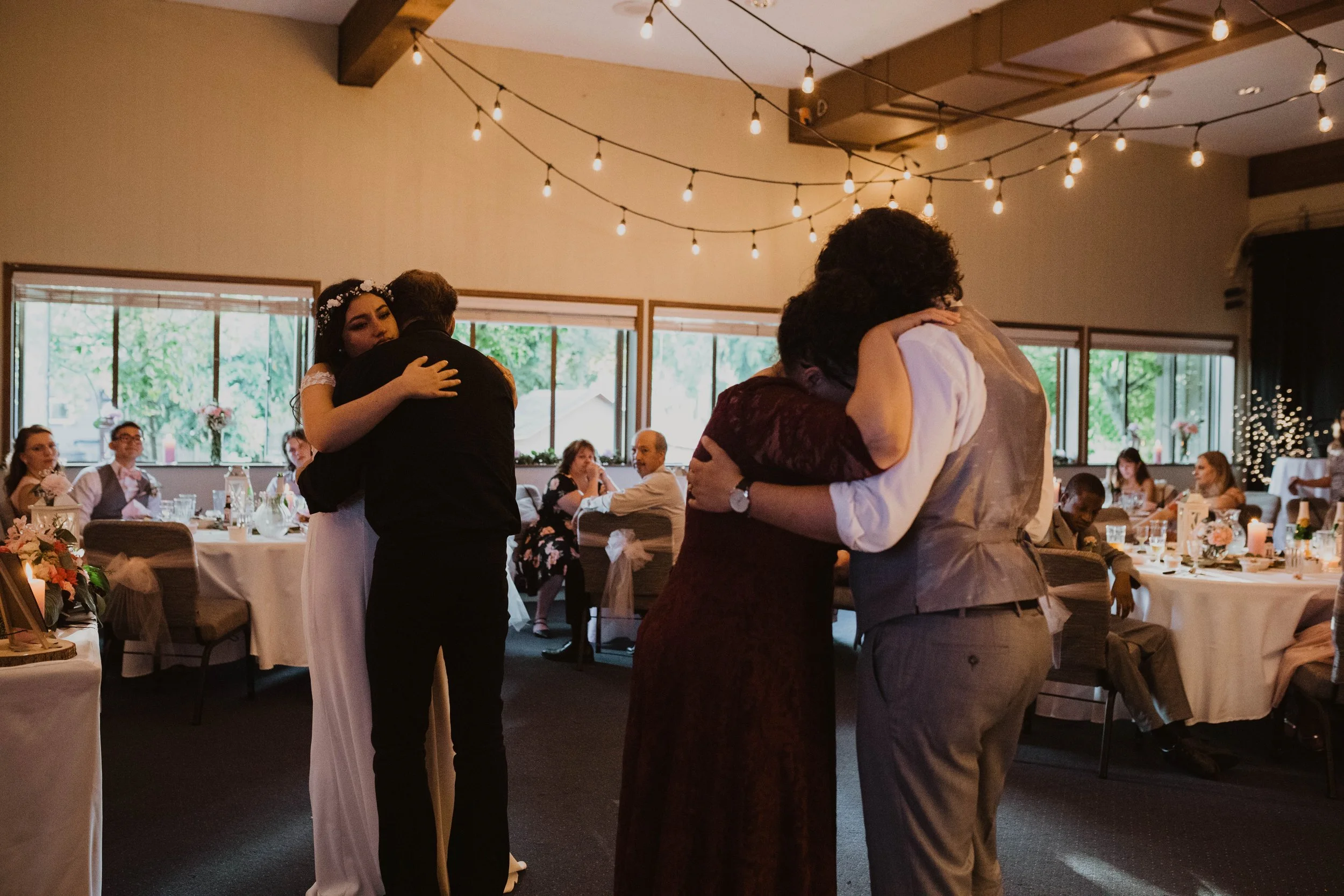 Two couples are embracing and dancing at a wedding reception in a decorated indoor venue with string lights and large windows showing greenery outside. Seattle, WA wedding photography.