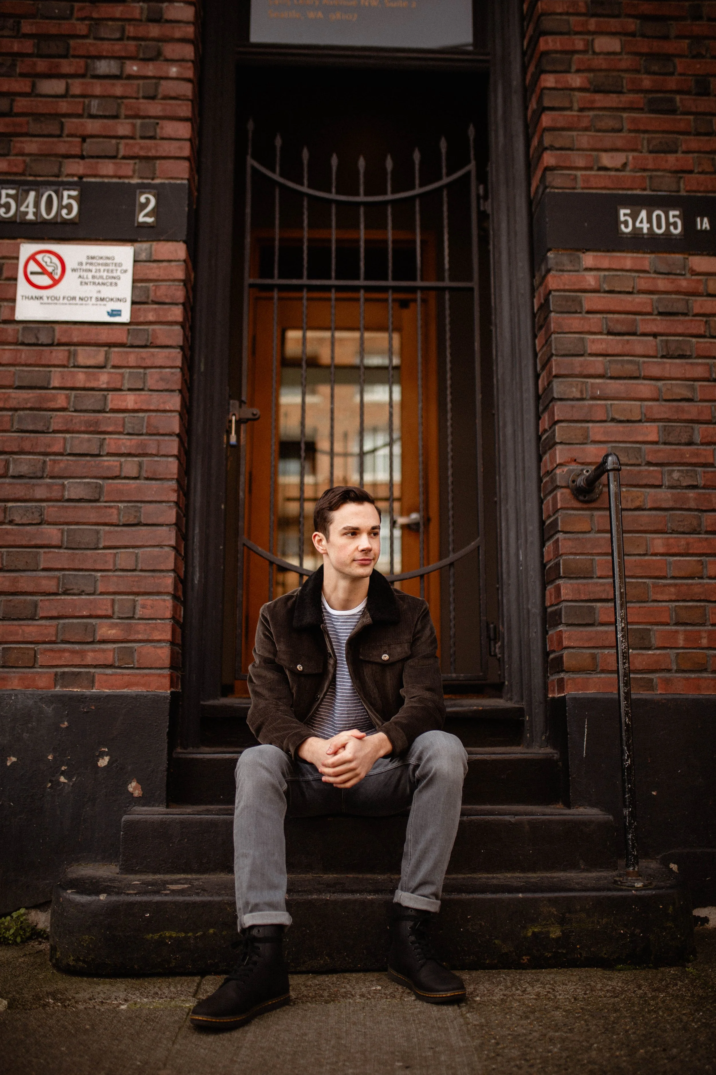 A young man sitting on the steps in front of a brick building with a metal gate and door behind him, wearing a brown jacket, striped shirt, gray jeans, and black boots. Seattle professional head shot photography