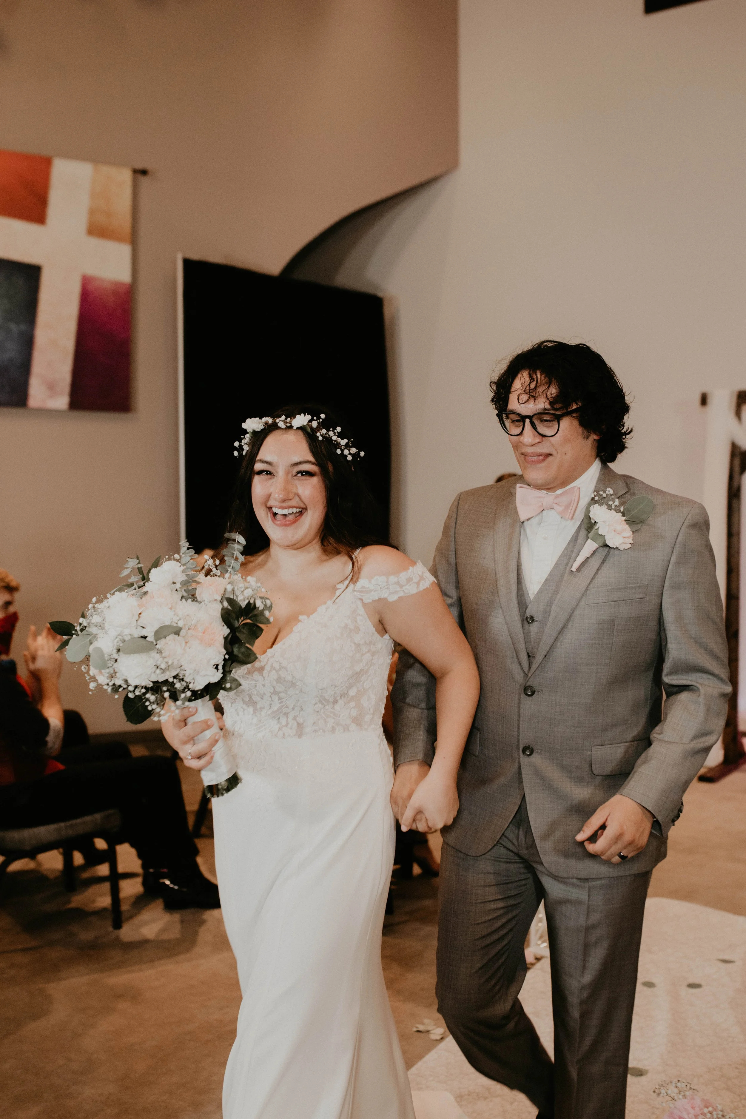 A smiling bride in a white lace wedding dress holding a bouquet of flowers walks hand in hand with a groom in a gray suit with a pink bow tie, at a wedding reception. Seattle, WA wedding photography.