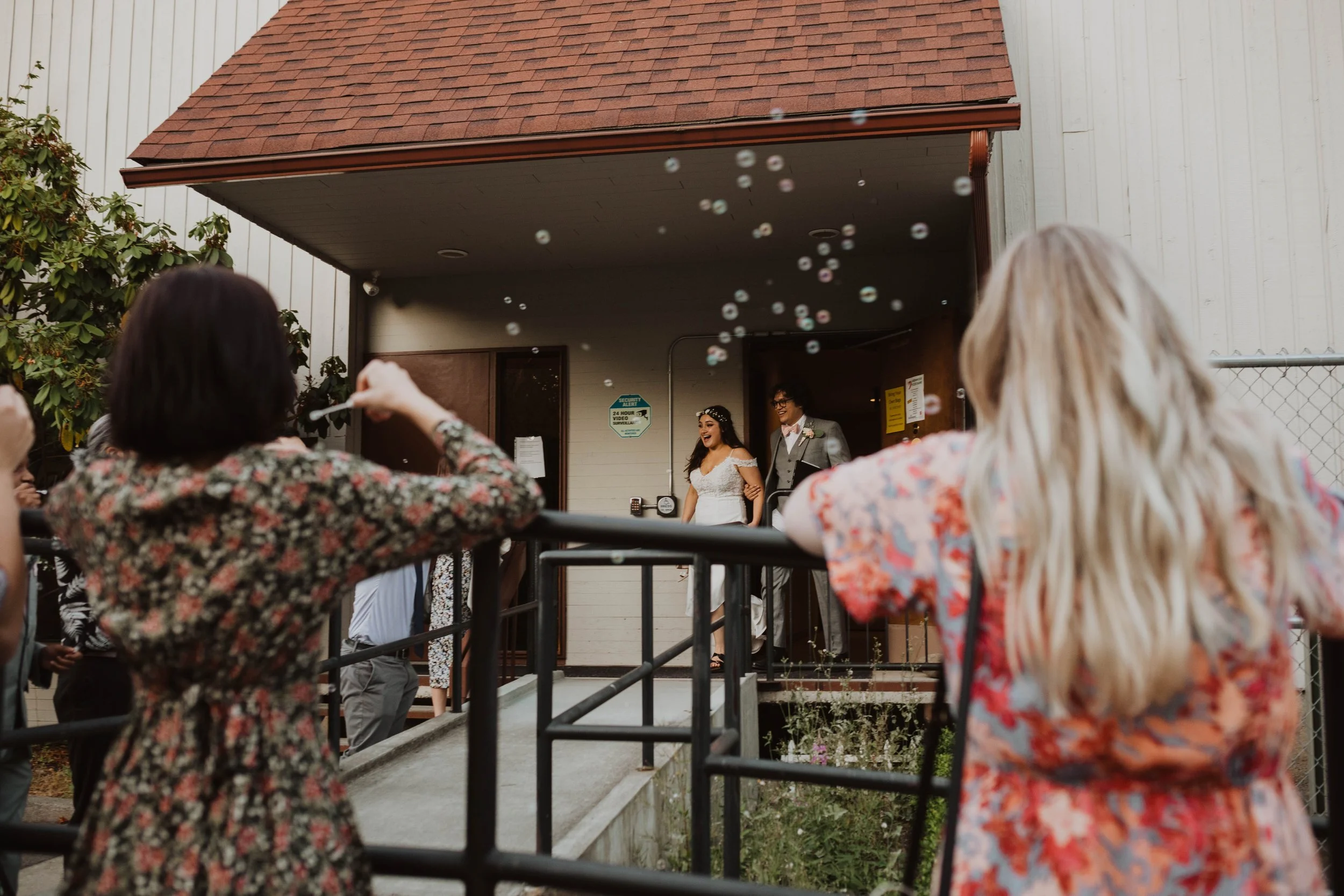 A bride and groom standing on a porch, smiling and holding hands, as guests celebrate with bubbles in the air. Seattle, WA wedding photography.