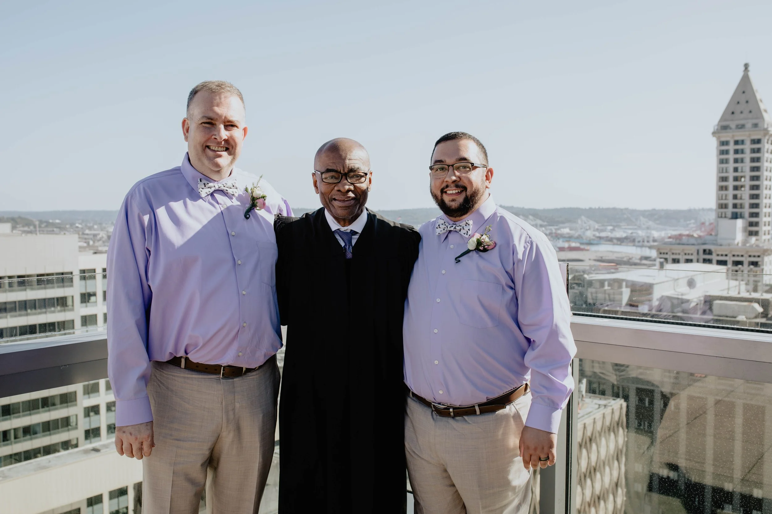 Three men dressed in wedding attire, standing on a balcony with a cityscape and water in the background, smiling at the camera. Seattle Municipal Courthouse wedding photography.
