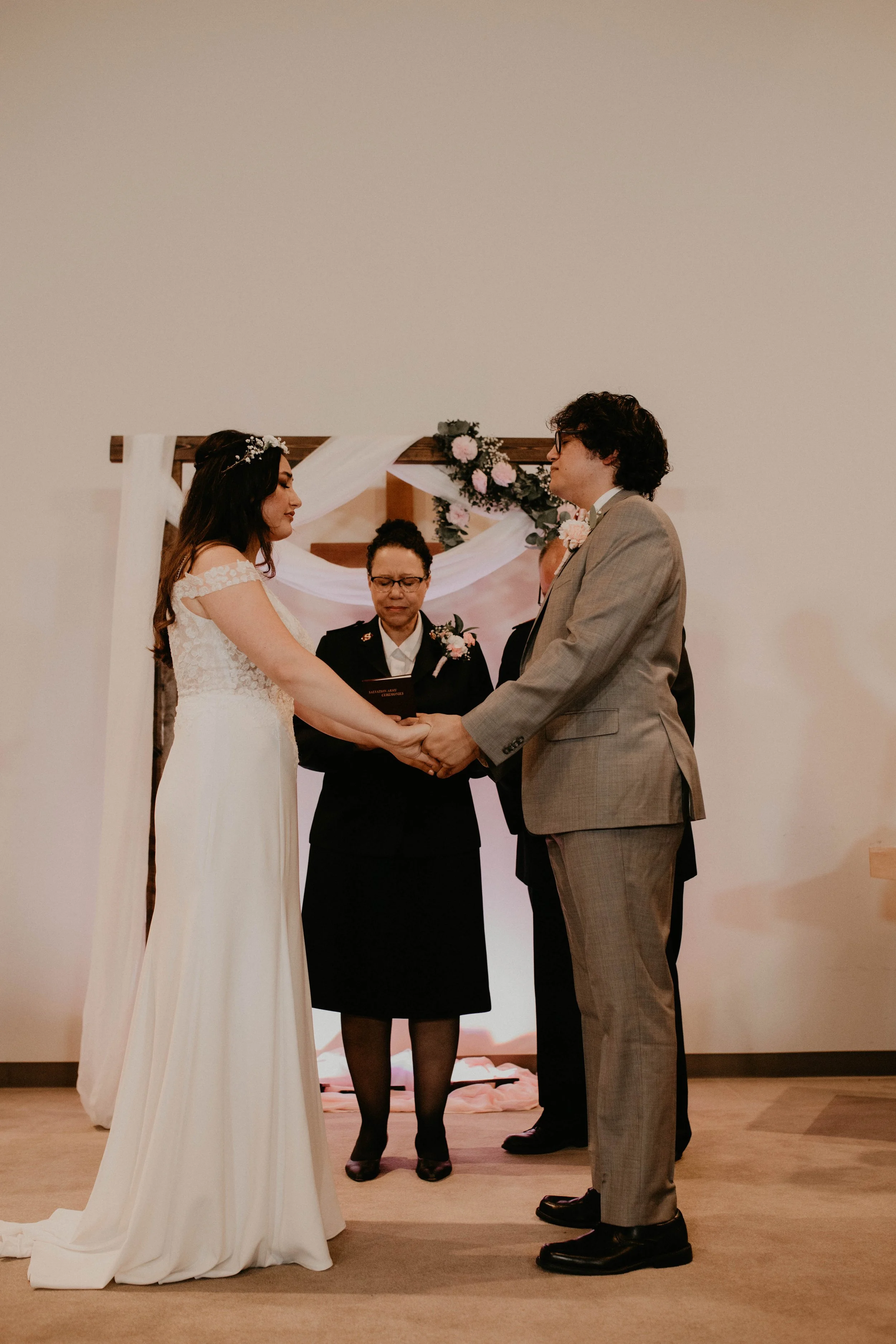 A couple getting married, holding hands and facing each other, with an officiant standing behind them during their wedding ceremony, decorated with pink and white flowers. Seattle, WA wedding photography.