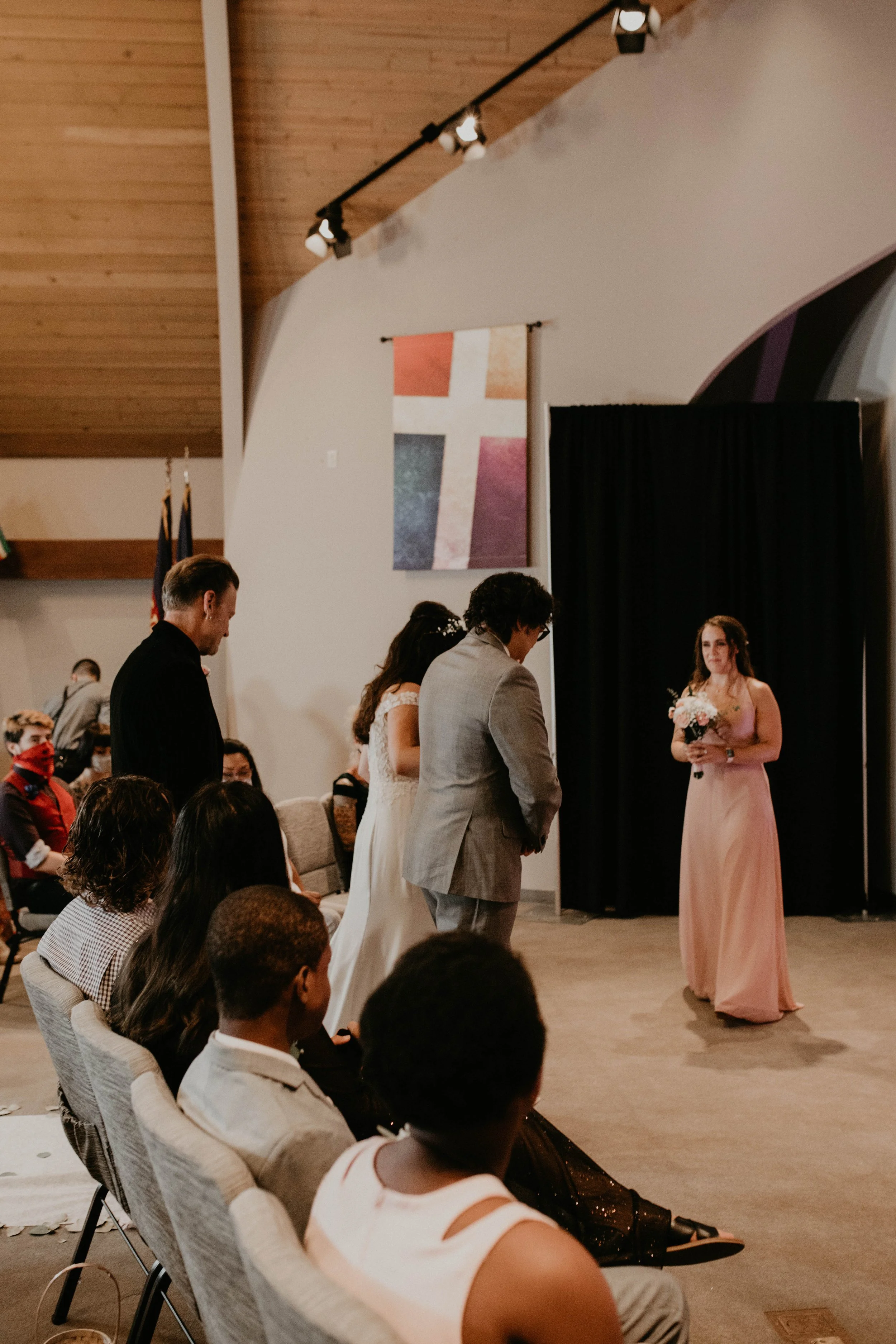 A wedding ceremony with a couple standing before a woman holding a bouquet on a stage. Guests seated and standing watch as the couple stands with heads bowed. The setting has a wooden ceiling and a large cross mural hanging on the wall.