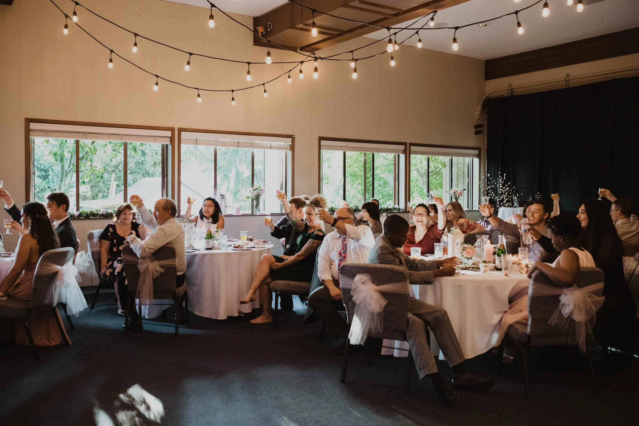 People celebrating at a wedding reception, sitting at round tables with white tablecloths and decorated chairs, raising glasses in a toast. The room has large windows, hanging string lights overhead, and some floral centerpieces on the tables.