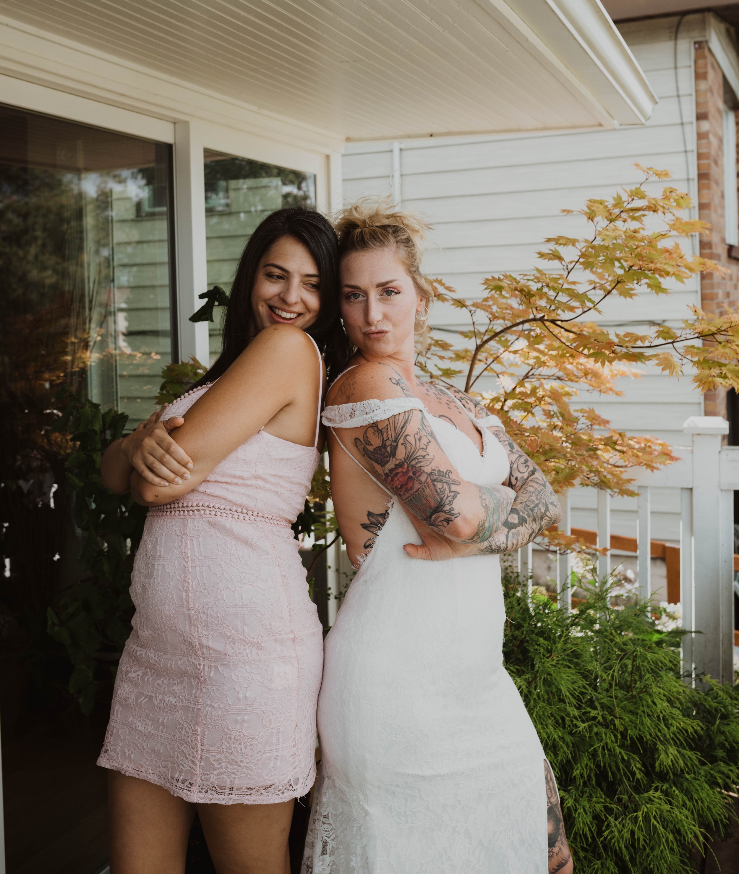 Two women stand close together outside, smiling and posing for a photo. One woman is wearing a pink lace dress, and the other is in a white dress with tattoos on her arms. There is foliage and a house in the background.
