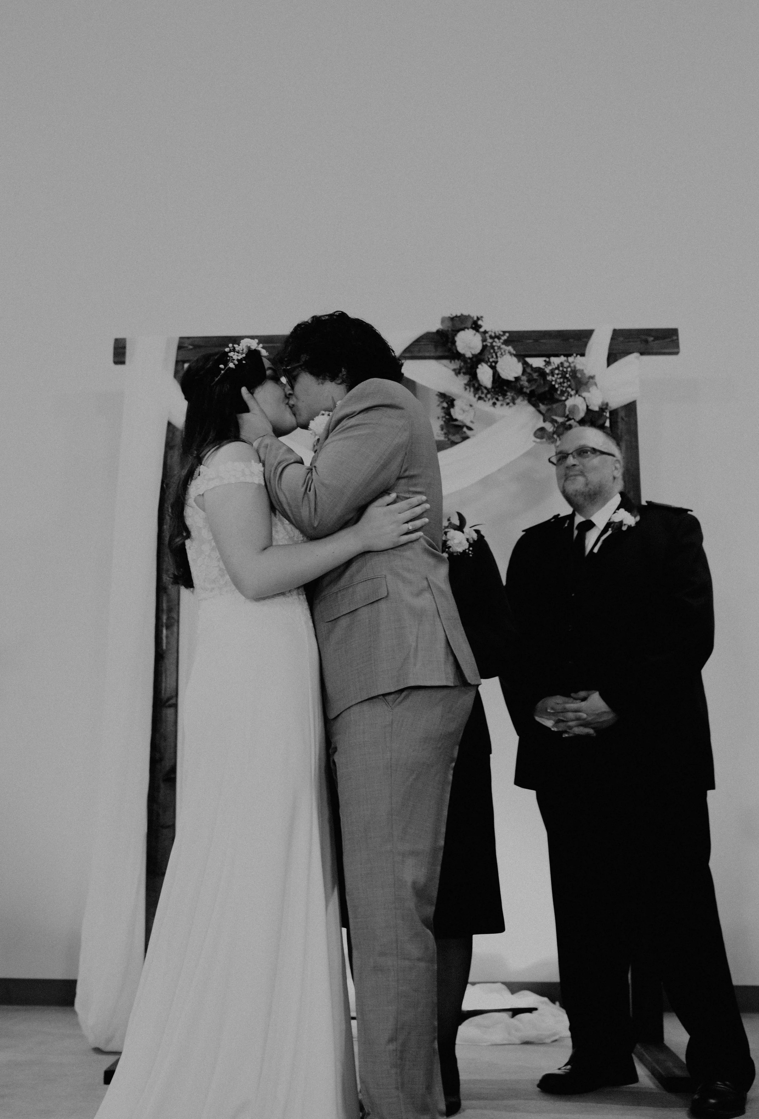 A couple sharing a kiss during a wedding ceremony with a officiant standing nearby in front of a floral arch. Seattle, WA wedding photography.